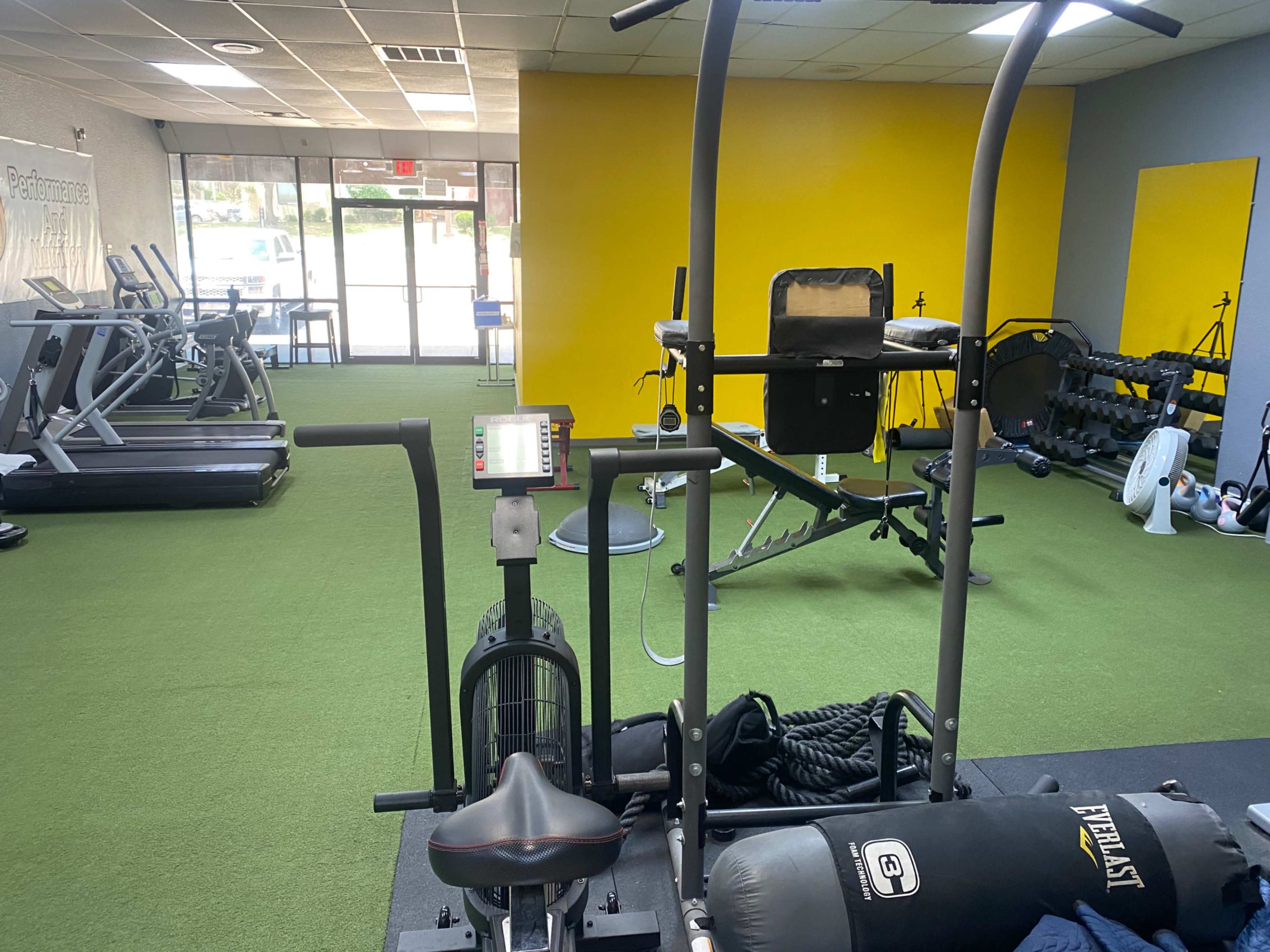 A gym interior with various exercise equipment, including treadmills, a rowing machine, a weight bench, and free weights, set against a yellow wall.