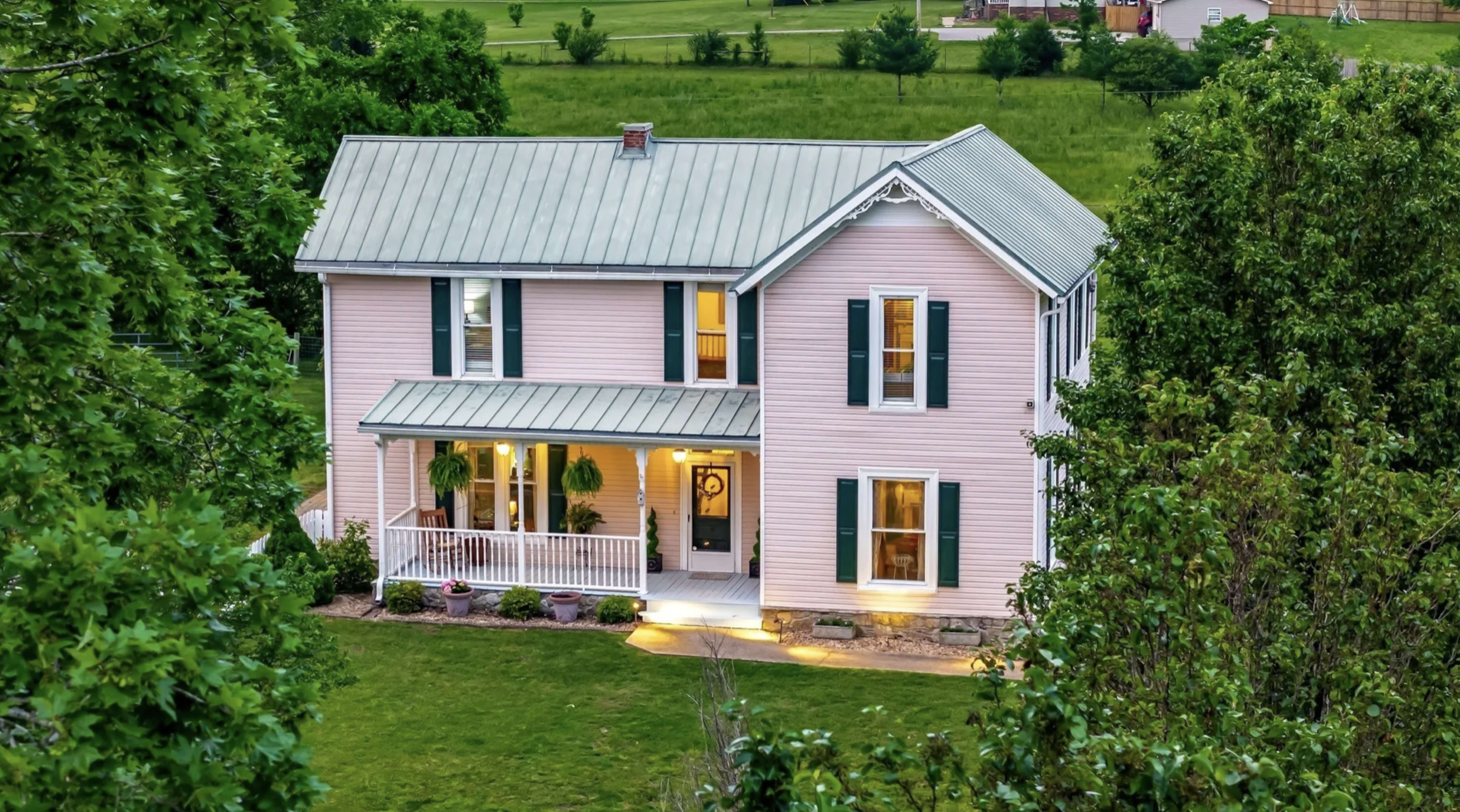 A two-story pink house with a metal roof and green shutters is surrounded by greenery and features a porch with lights on at dusk.