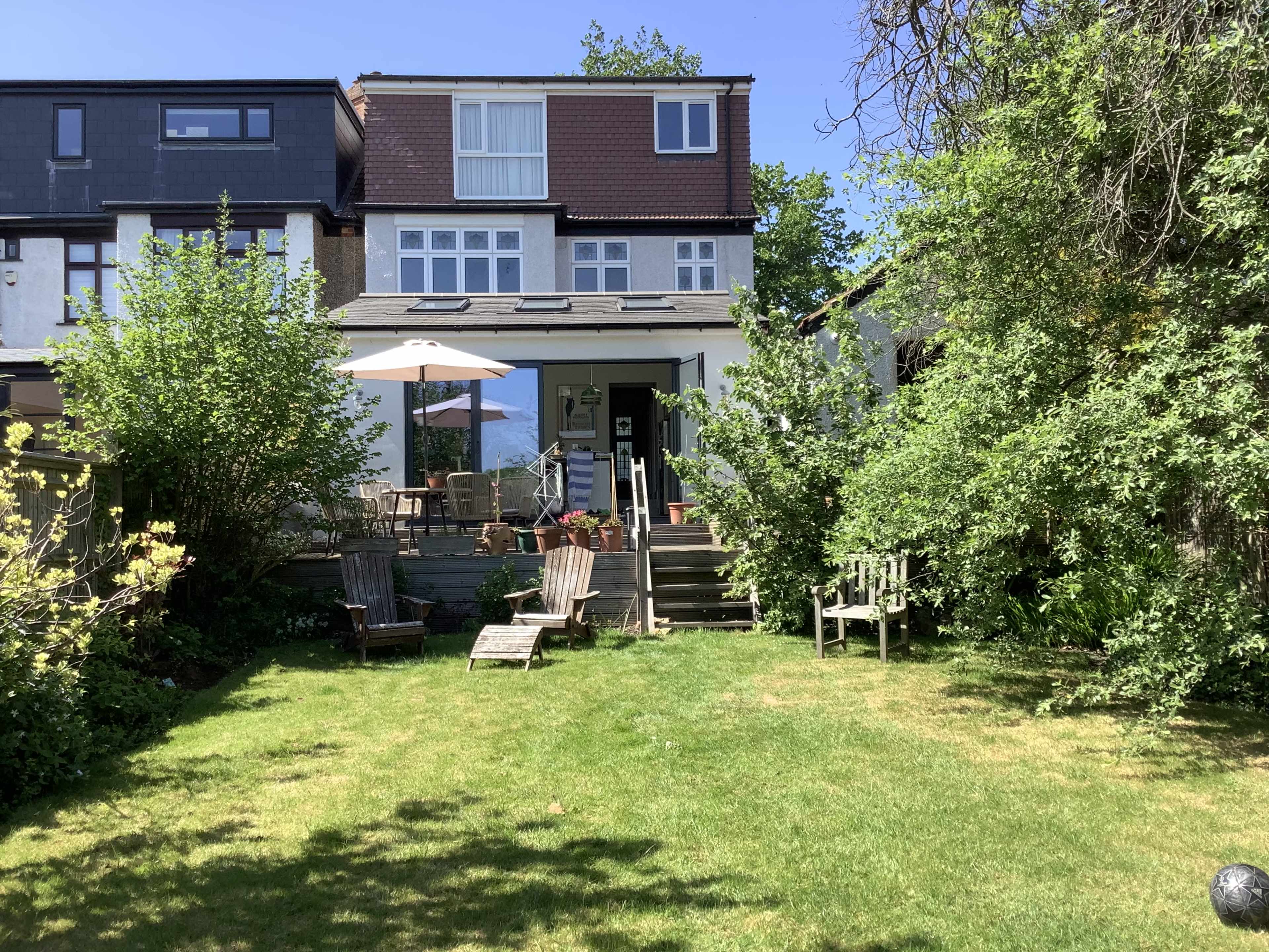 A two-story house with a patio sits at the back of a green backyard, surrounded by trees and lawn chairs.