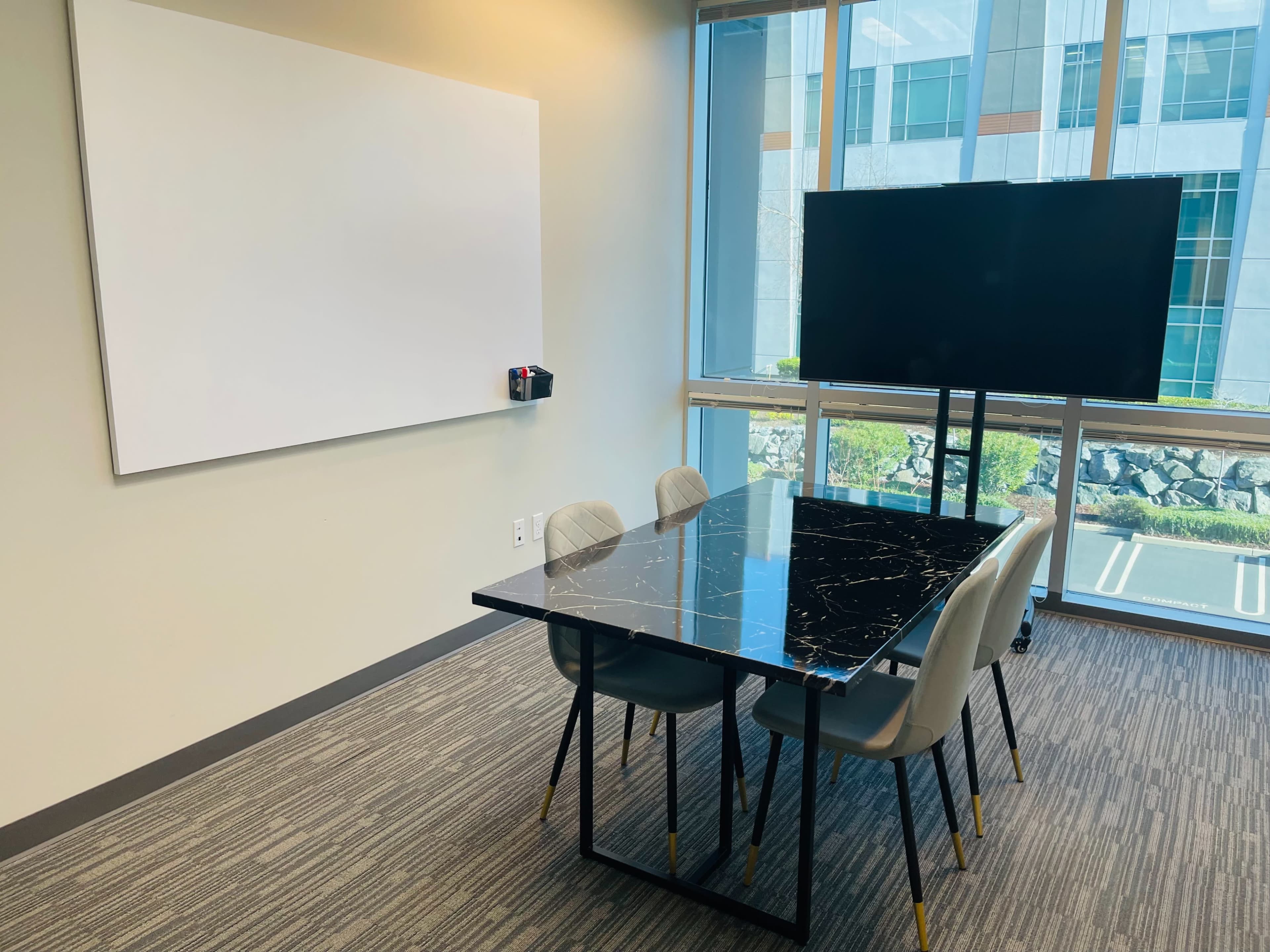 A modern conference room features a black marble table, four gray chairs, a whiteboard on the wall, and a television mounted on a stand.