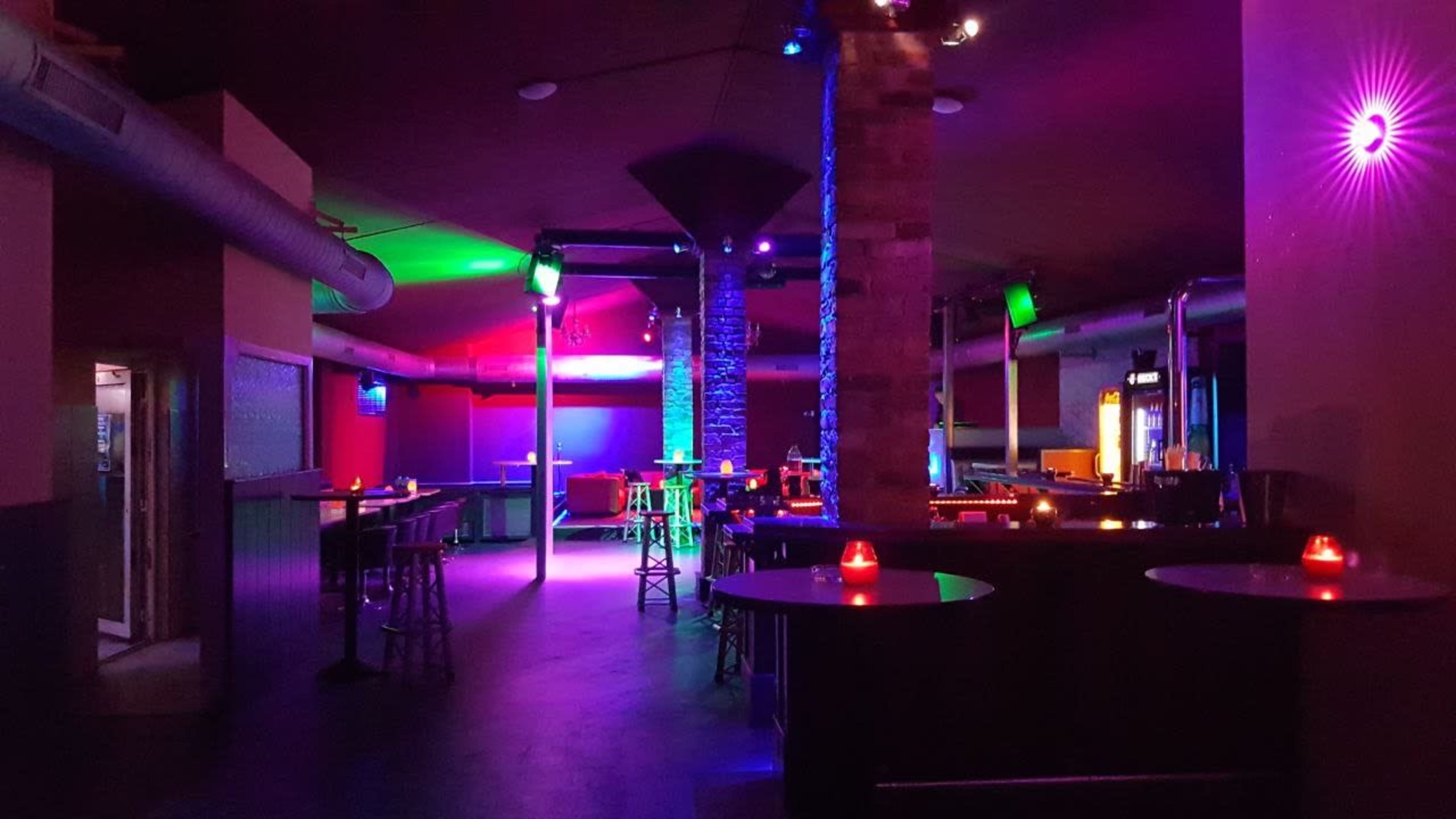 The image shows a dimly lit bar with colorful lighting, featuring high tables, stools, and a few scattered candles on the tables.