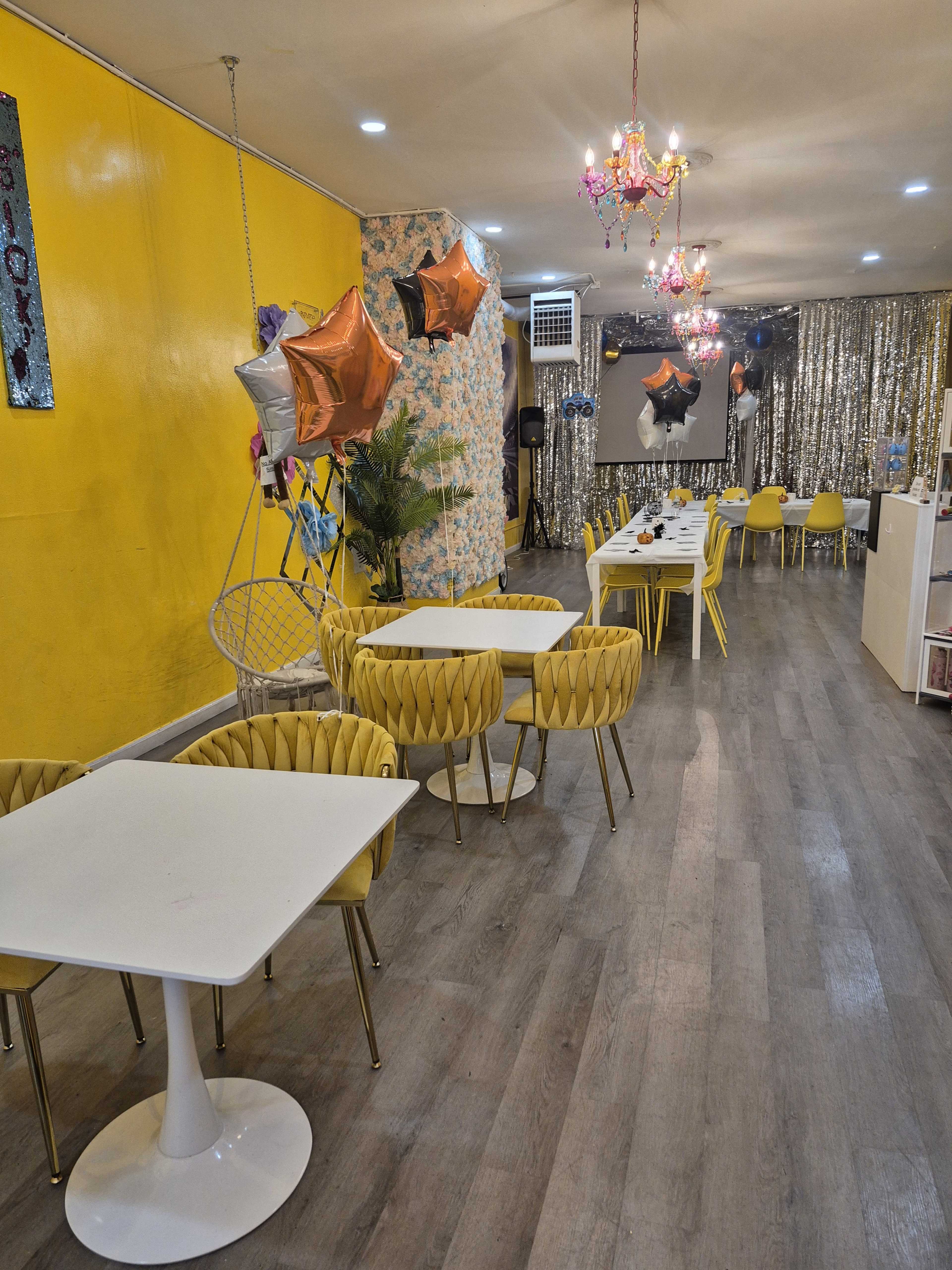 The image shows a brightly colored café with yellow walls, gold-accented chairs, and a glittery backdrop, set for a celebration with balloons and decorative lighting.