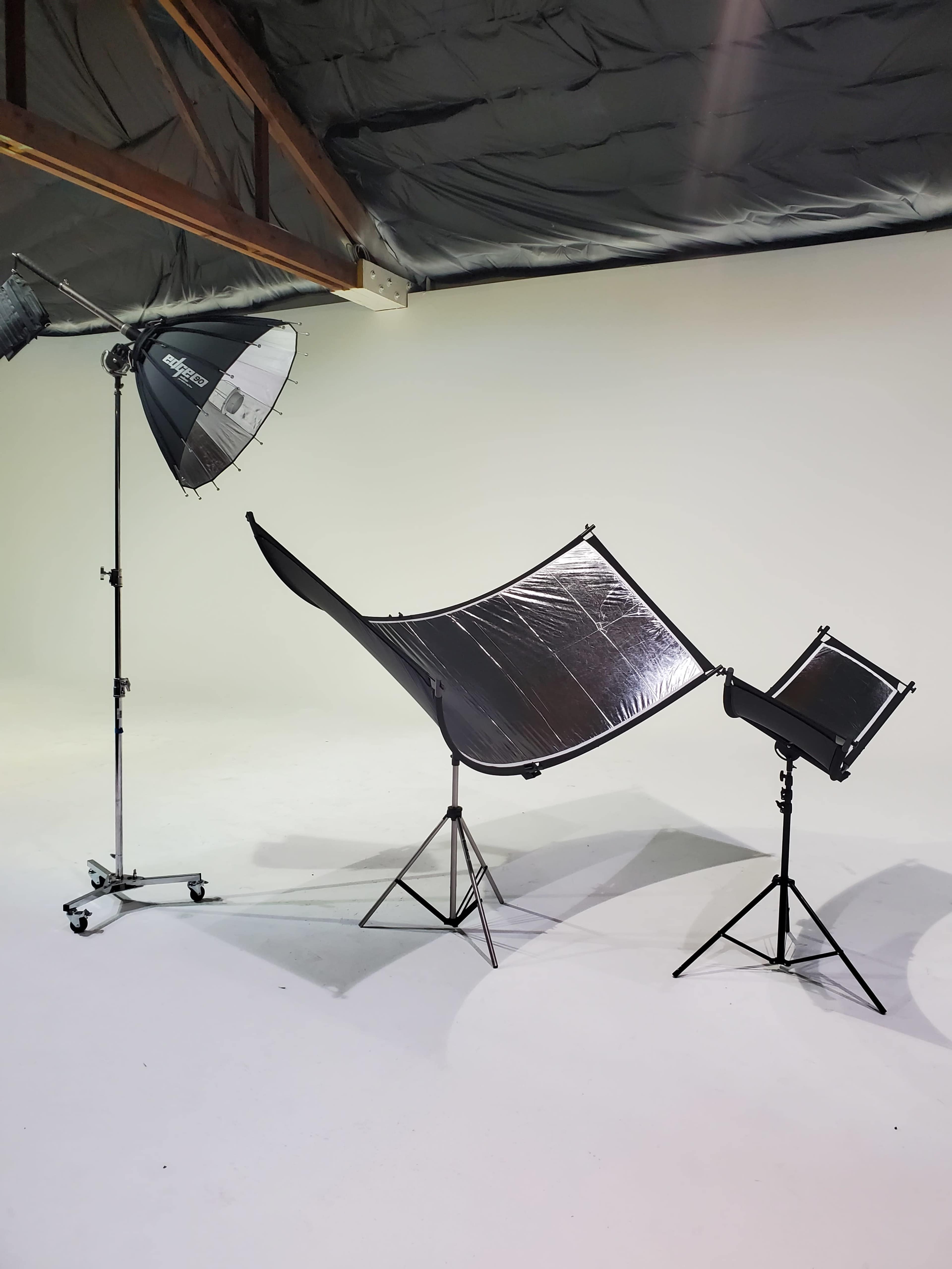 A photography studio setup featuring a large softbox light, a curved reflector, and a smaller light setup on stands against a plain background.
