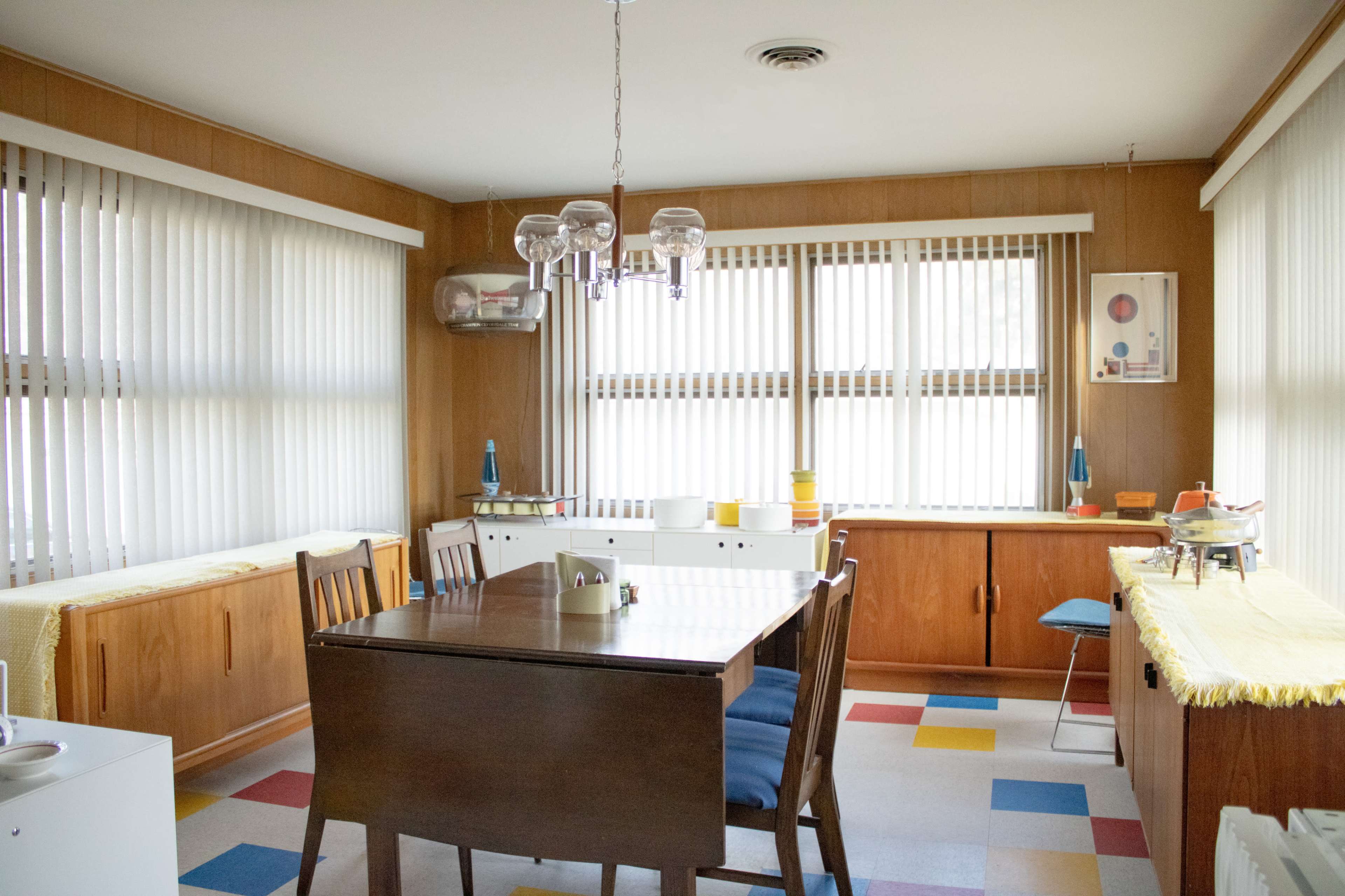 A well-lit dining area features a wooden table surrounded by chairs, with colorful tiled flooring and large windows covered by vertical blinds.