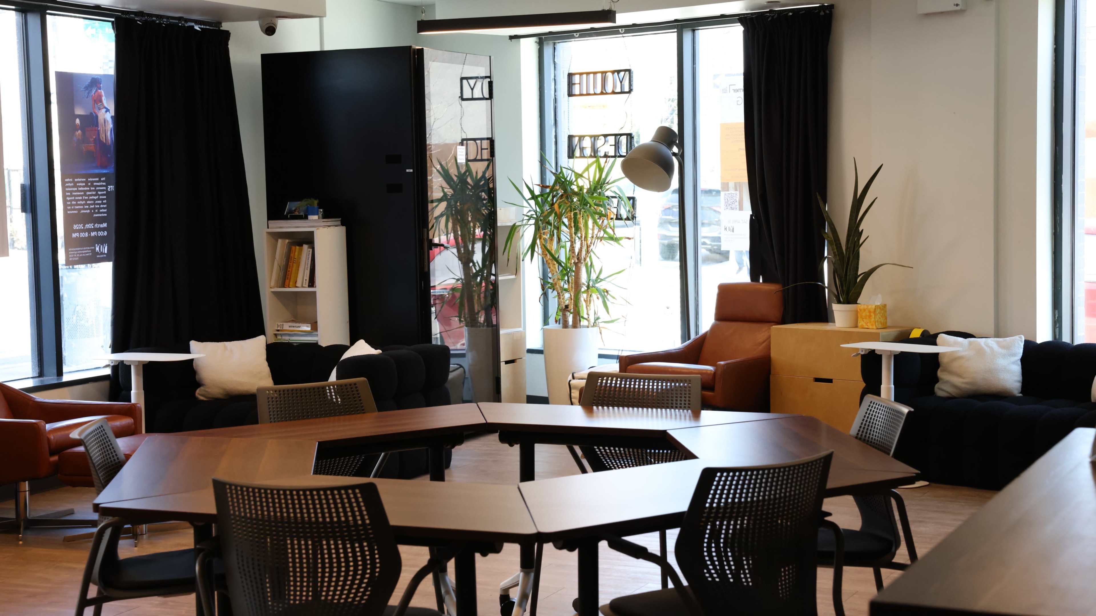The image shows a modern common area with a hexagonal wooden table surrounded by sleek chairs, plants, and a mix of seating options including a black couch and an orange armchair near large windows.