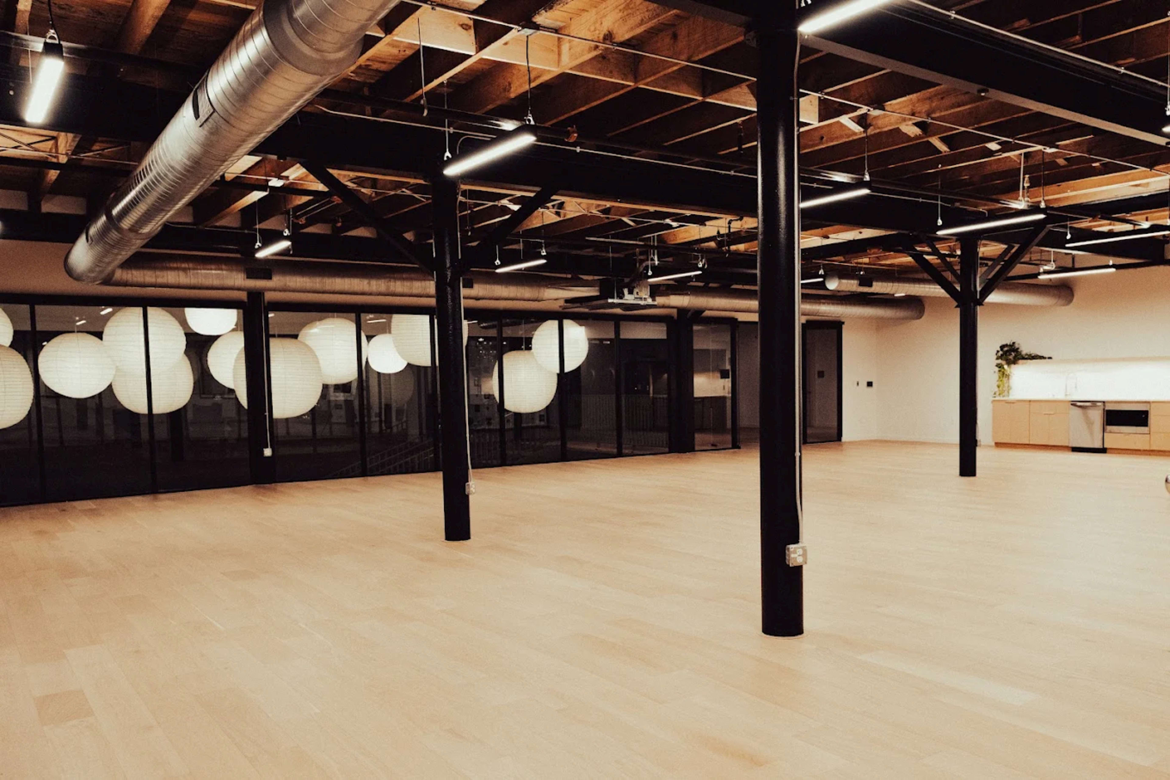 The image shows an empty, spacious interior with wooden flooring, exposed beams, and large windows featuring round light fixtures.