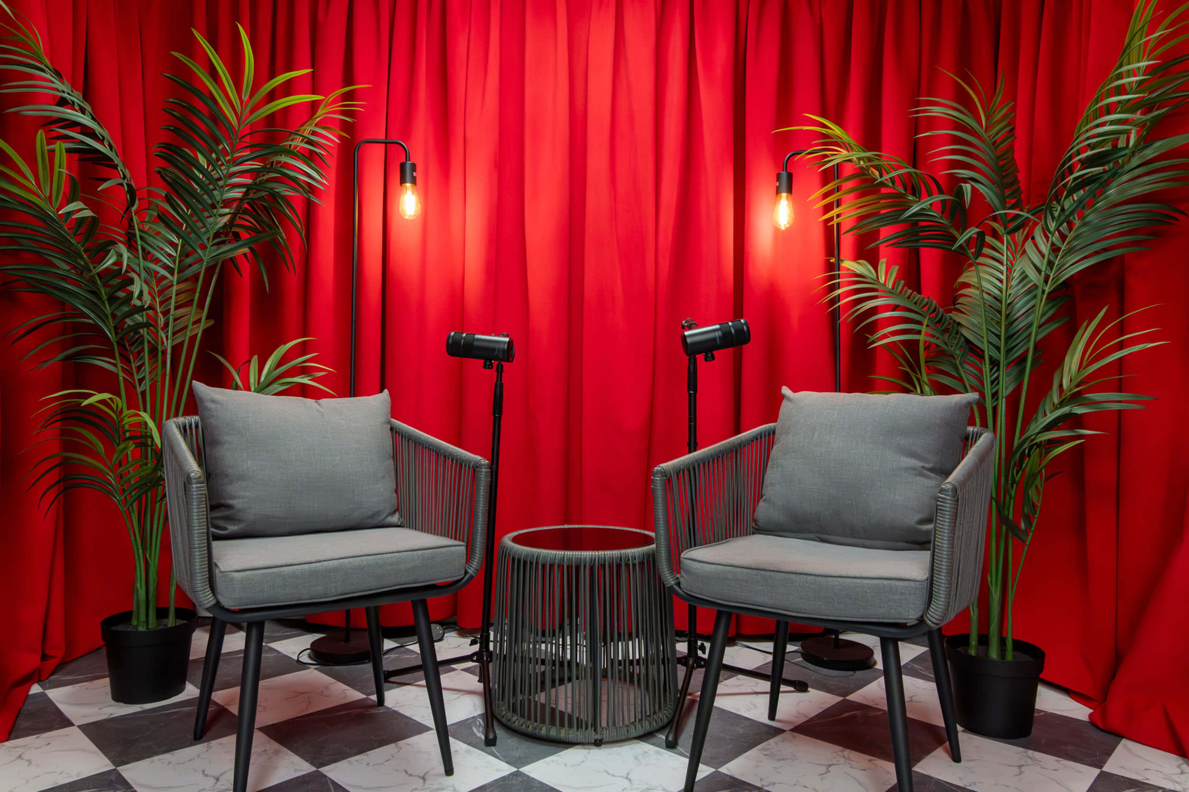 A pair of gray chairs and a round table are positioned in front of a bright red curtain, flanked by plants and studio lights.
