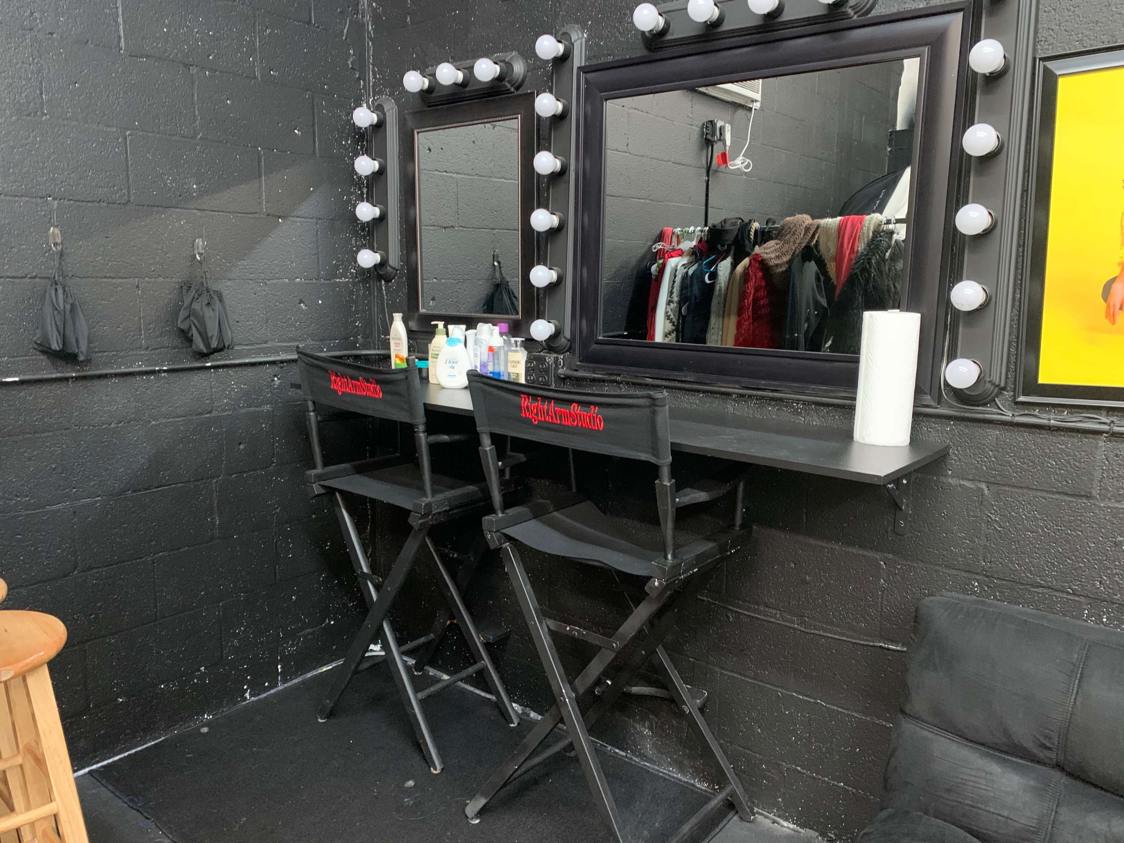 The image shows a black-walled room with two makeup chairs and a long mirror framed in black, flanked by illuminated vanity lights.