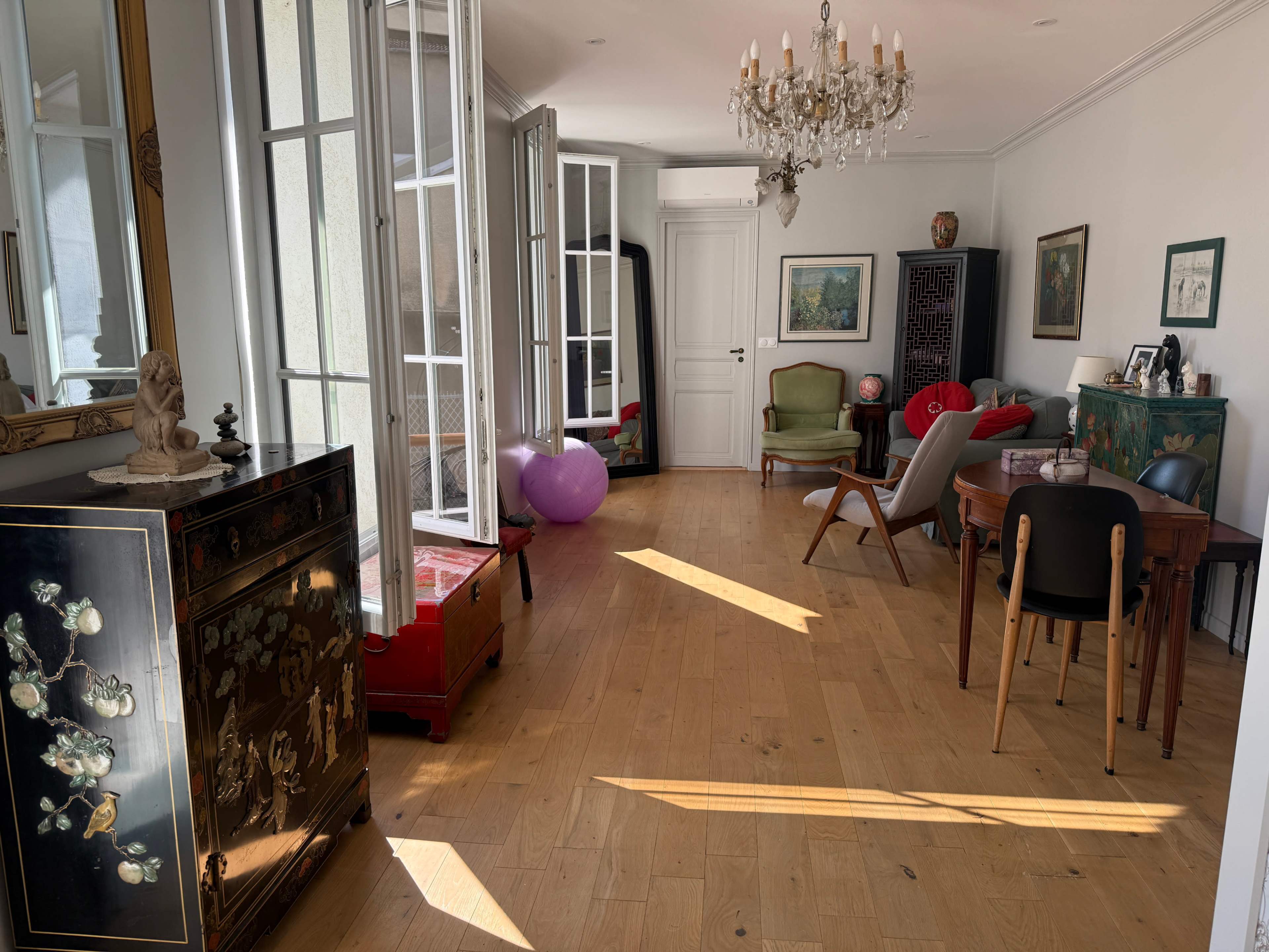 The image shows a brightly lit room featuring wooden flooring, a chandelier, various pieces of furniture including a dining table, chairs, and a decorative cabinet, with large windows allowing sunlight to enter.