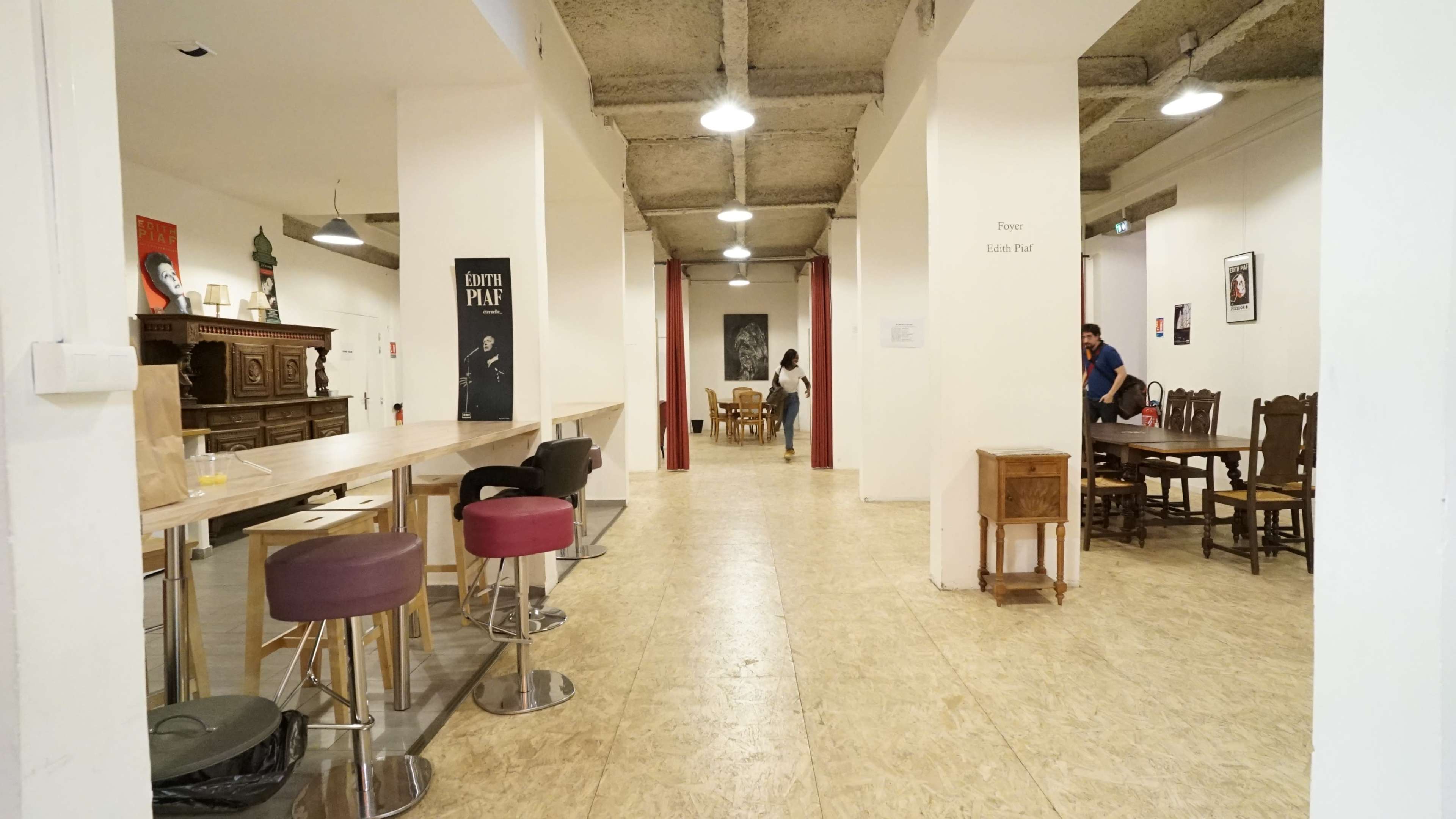 A spacious interior with a long table and stools on the left, wooden tables and chairs scattered throughout, and white walls with artwork displayed.