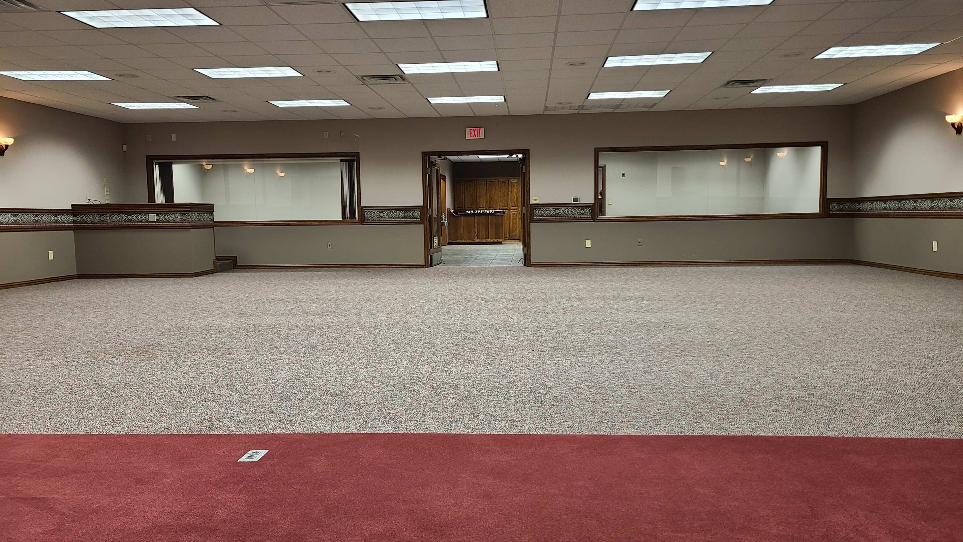 The image shows a spacious, empty carpeted room with a maroon section at the front and doorways leading to adjoining spaces.