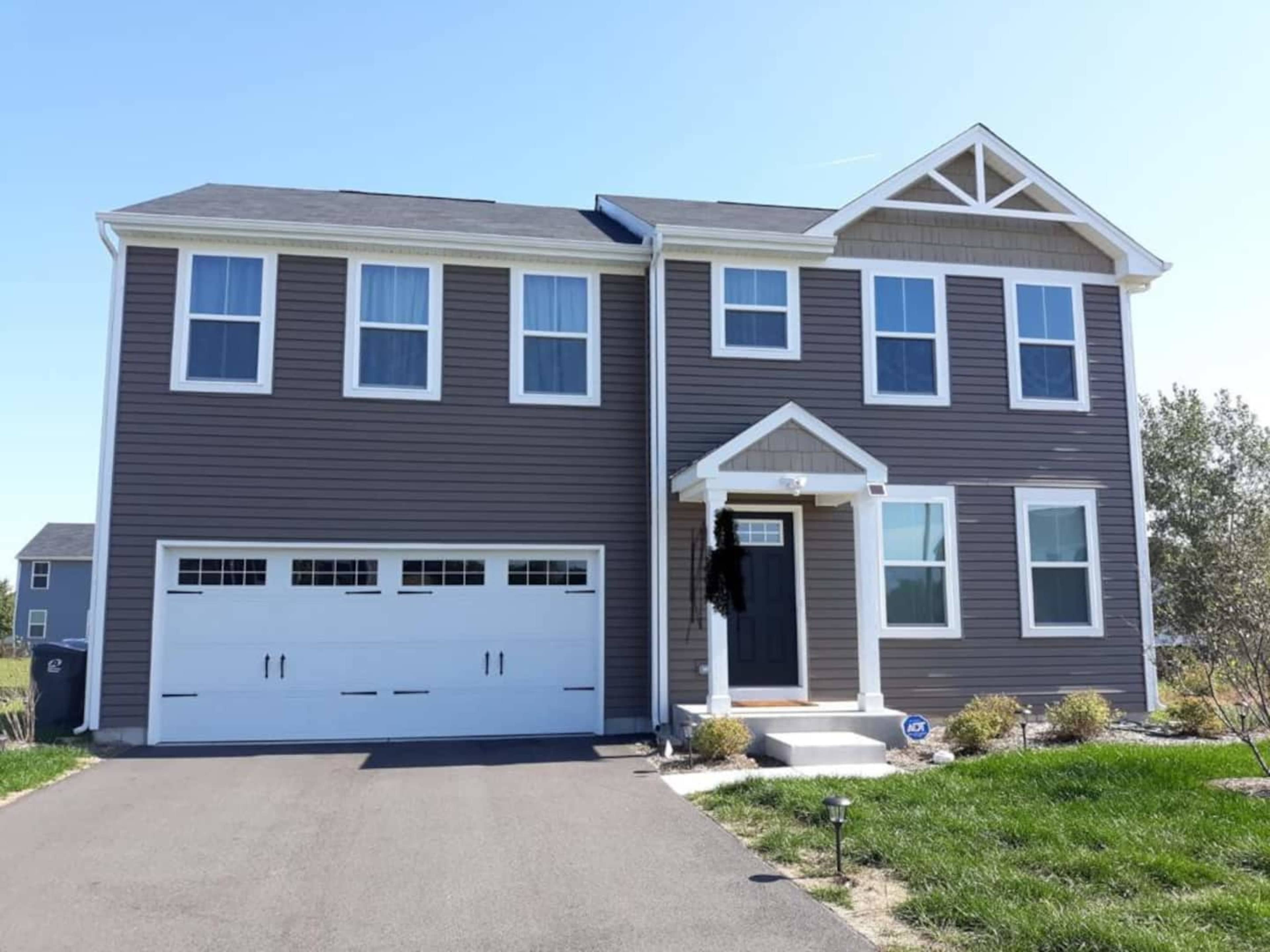 A two-story gray house with white trim and a front porch, featuring a garage and a well-maintained lawn.