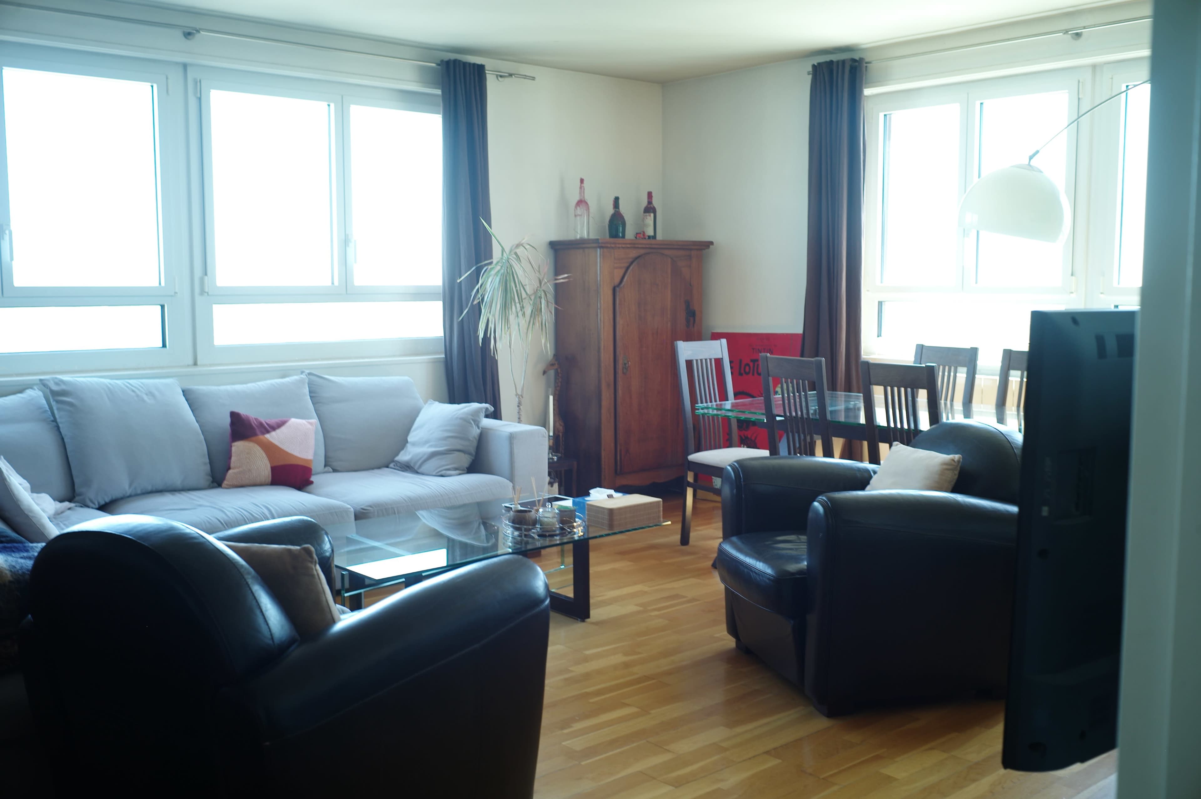 The image shows a bright living room furnished with a light-colored sofa, dark armchairs, a wooden dining table, and a wooden cabinet, all illuminated by natural light from large windows.