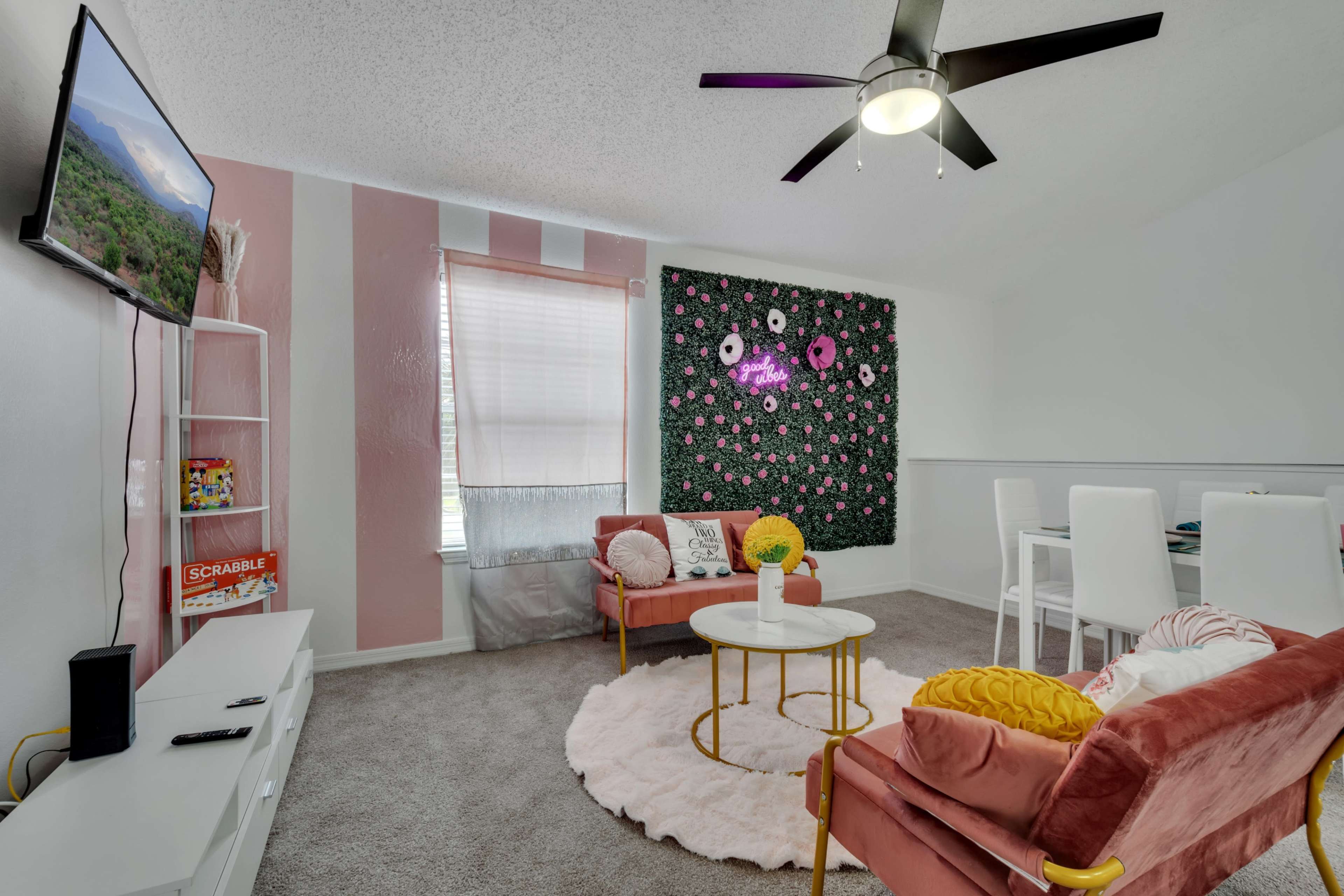 A cozy living space features pink-striped walls, a floral accent wall, two pink sofas, a round coffee table, and a dining area with white chairs.