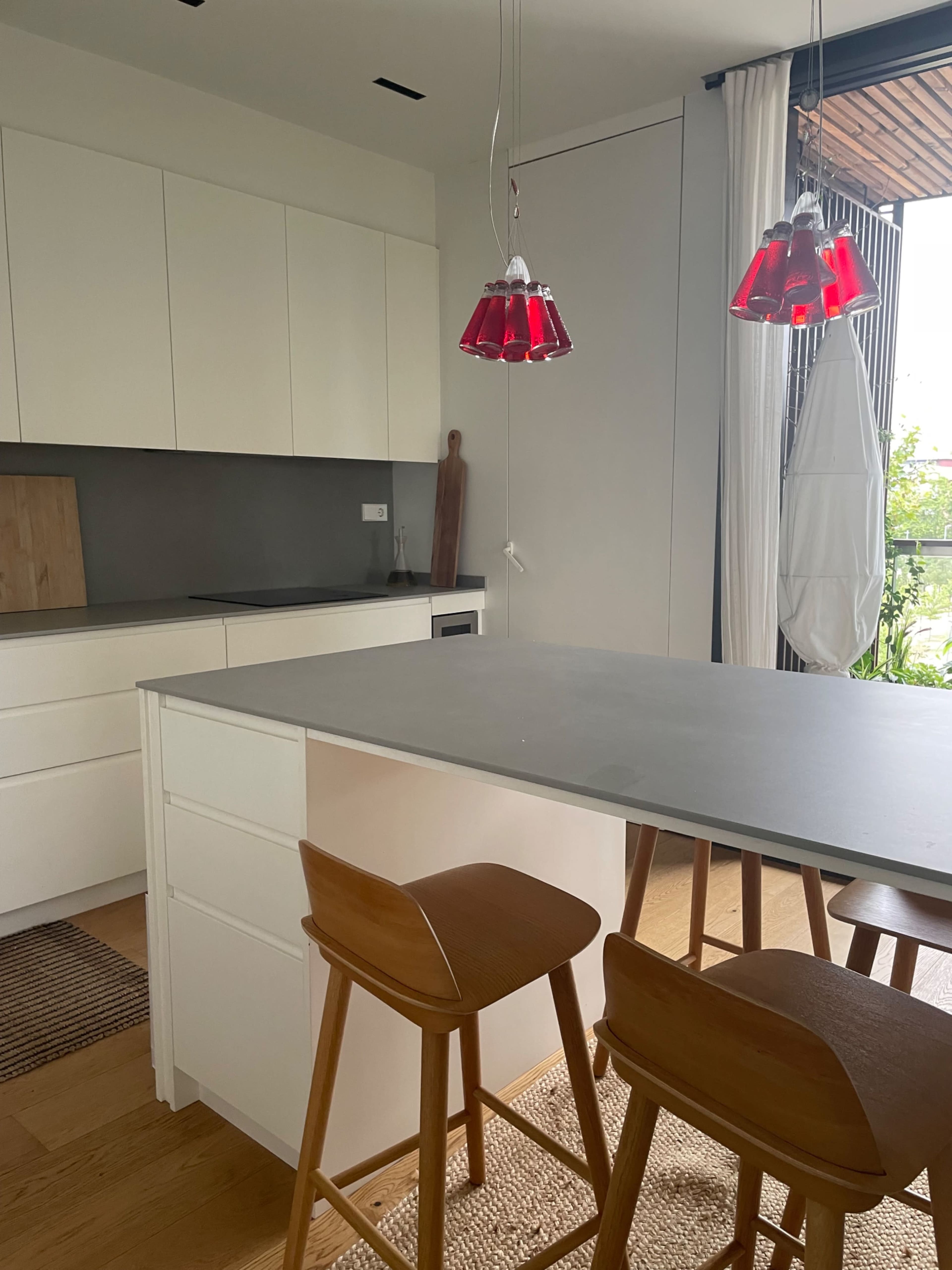 The image shows a modern kitchen with a central island, two wooden stools, and red pendant lights hanging above.