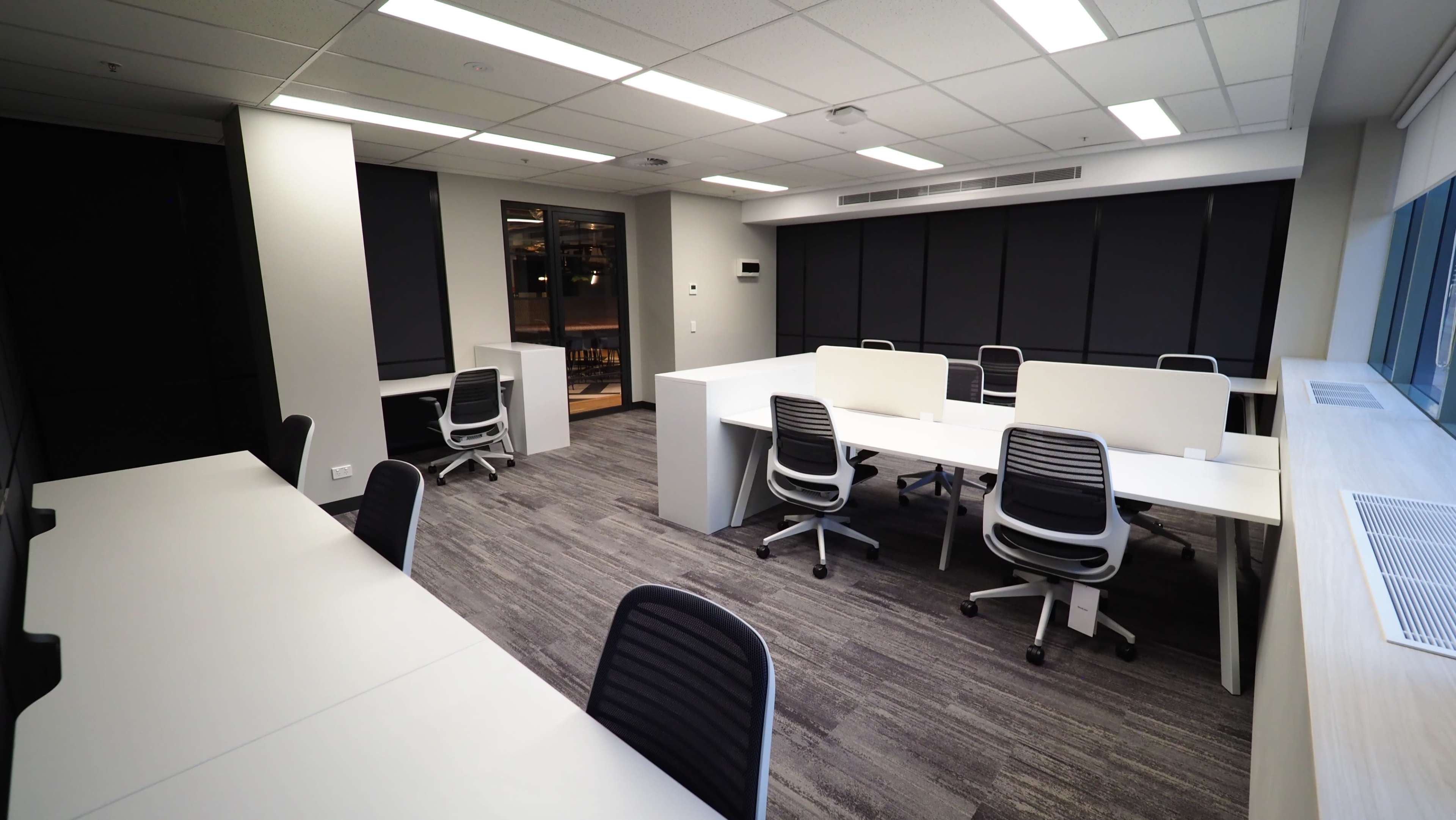 The image shows a modern office space with multiple workstations, each equipped with desks and ergonomic chairs, arranged in a well-lit layout.