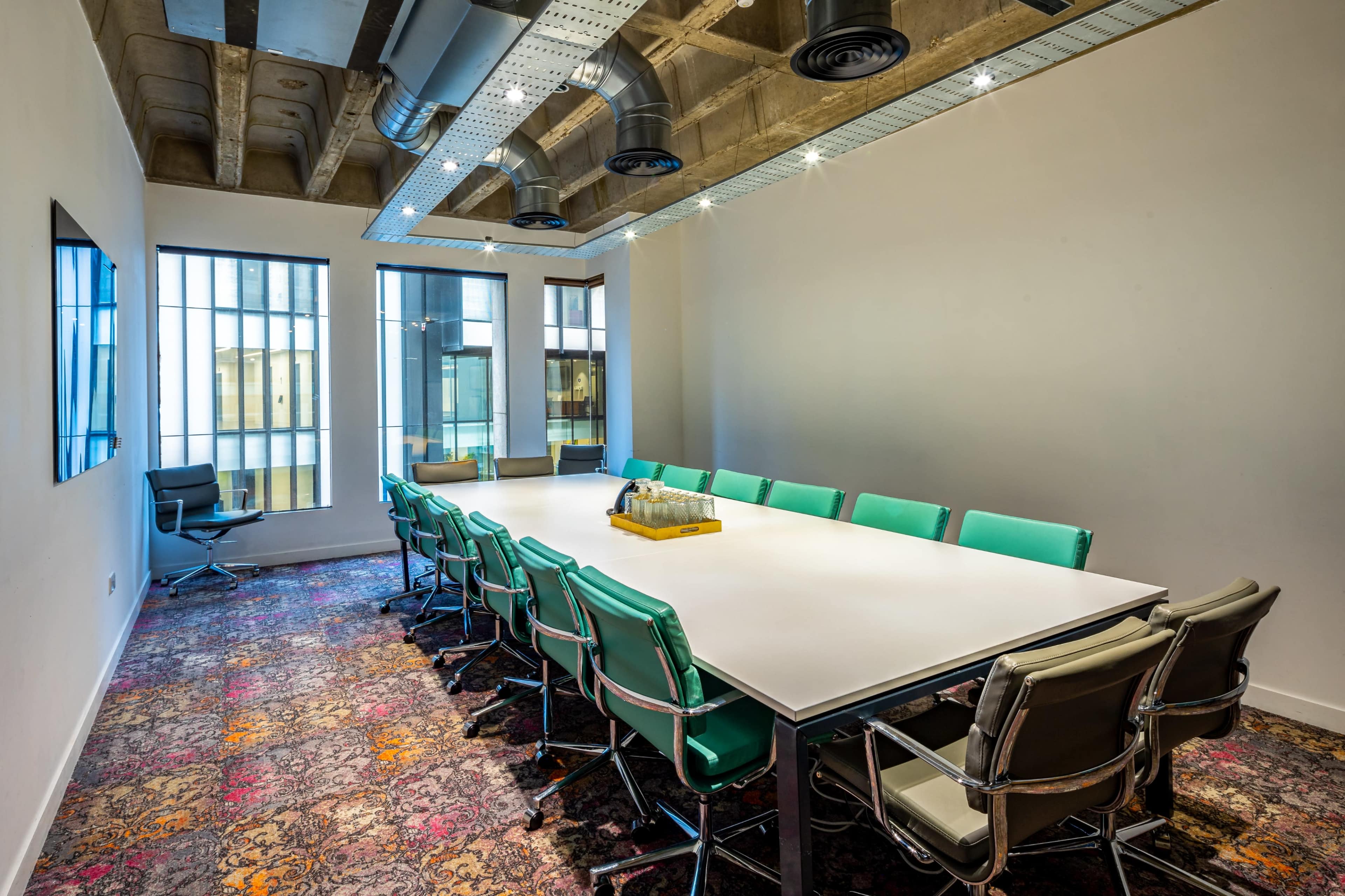 A large conference room features a rectangular table surrounded by turquoise and silver chairs, with large windows and exposed ceiling elements.