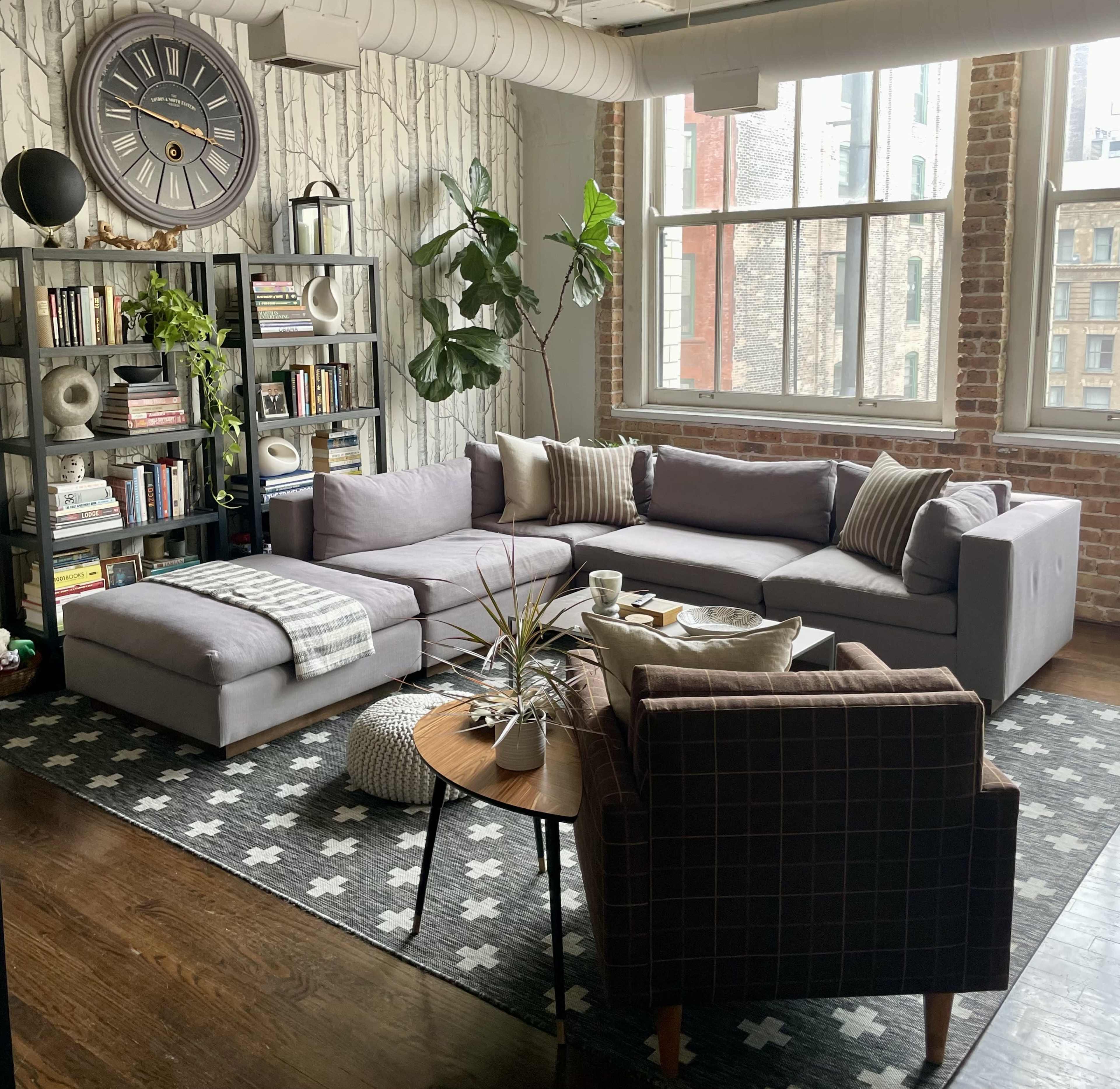 A modern living room features a gray sectional sofa, a round wooden coffee table, and a bookshelf filled with books, all set against a backdrop of brick walls and large windows.