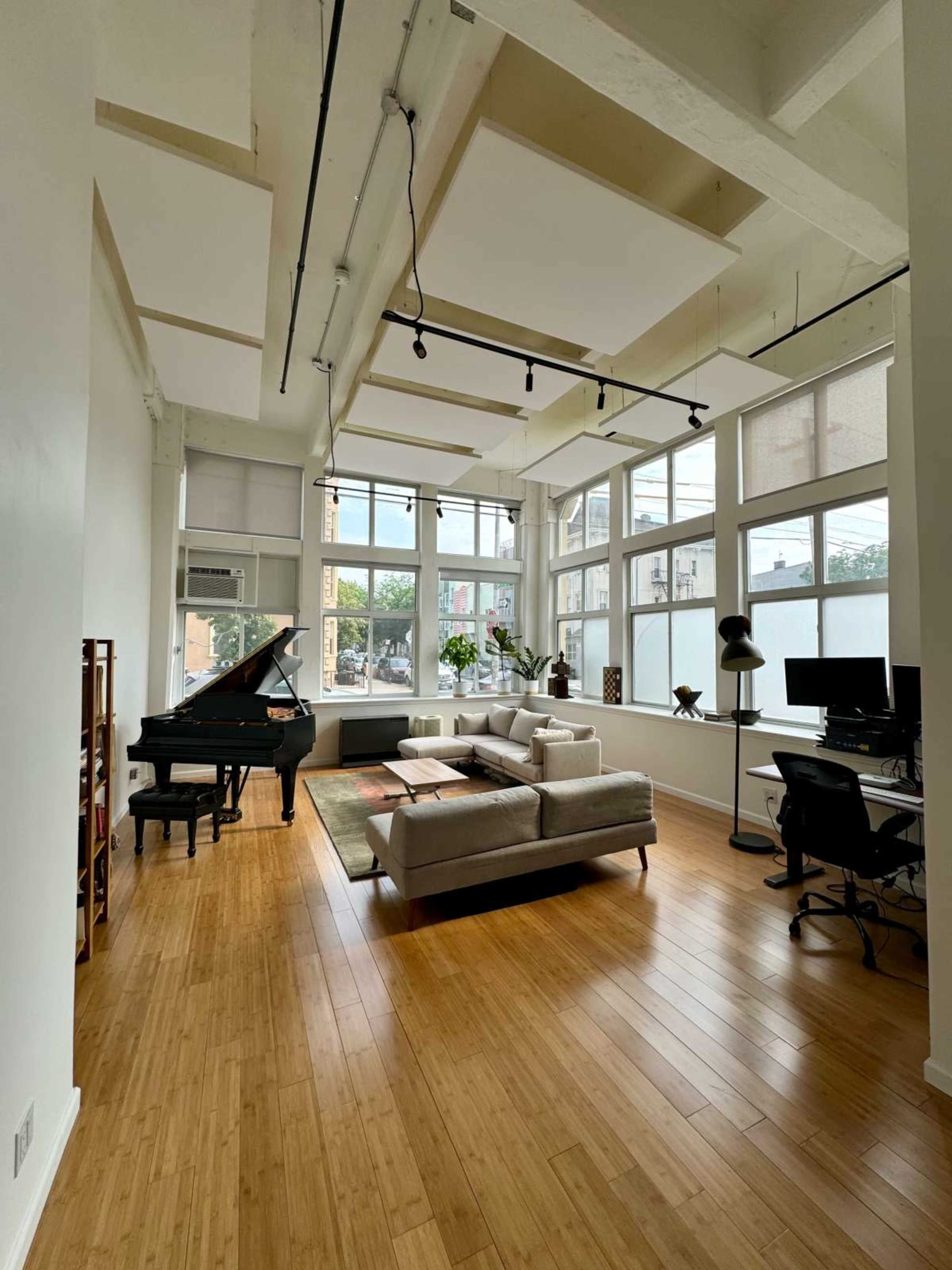 The image shows a spacious, modern living room with a grand piano, a sectional sofa, and a home office setup, featuring large windows that allow natural light to fill the space.