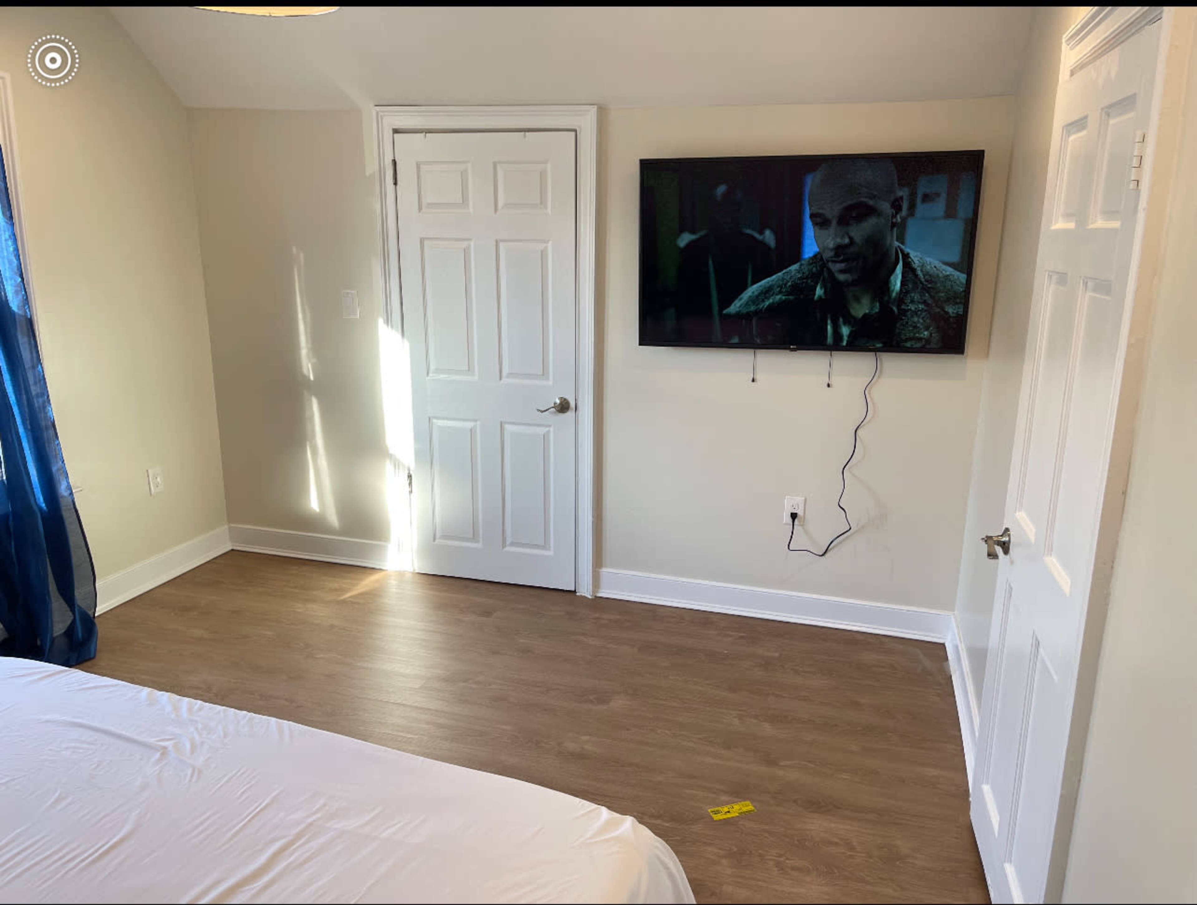 The image shows a sparsely furnished bedroom with a bed on one side, a wall-mounted television displaying a scene, and a door on the opposite wall.
