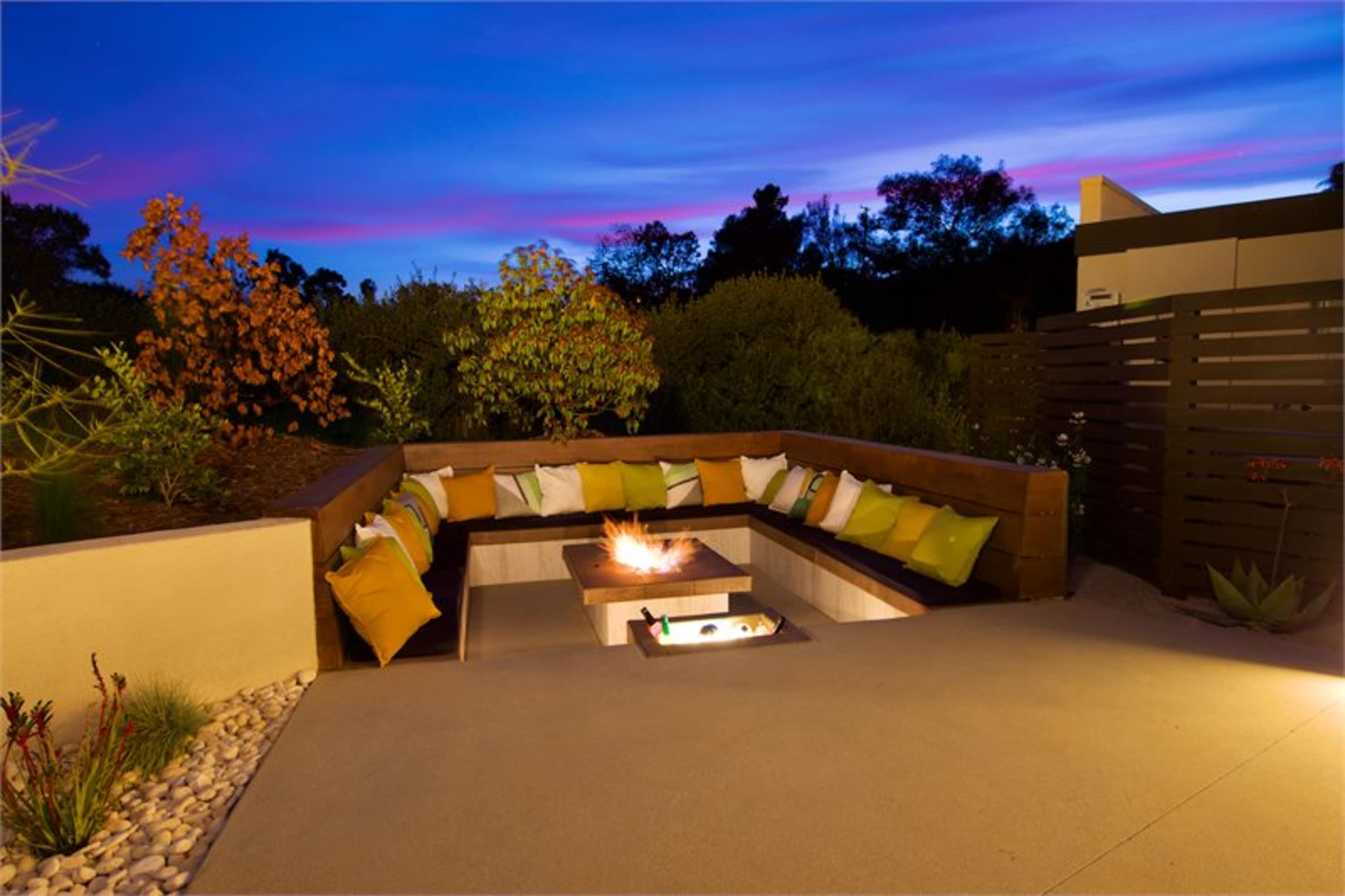 A modern outdoor seating area features a circular sofa with colorful cushions surrounding a central fire pit, set against a twilight sky.