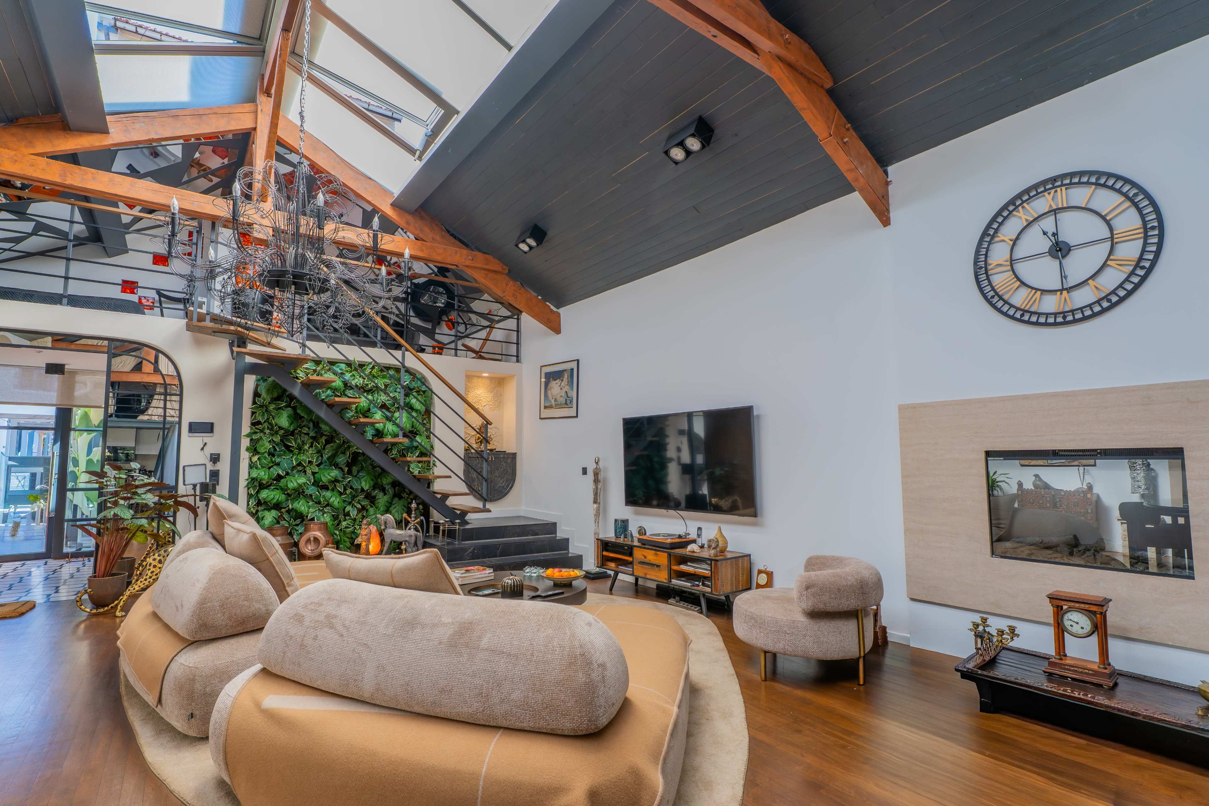 The image shows a modern living room with a high ceiling, large windows, a staircase, and a mix of furniture, including a curved sofa and a wall-mounted television.