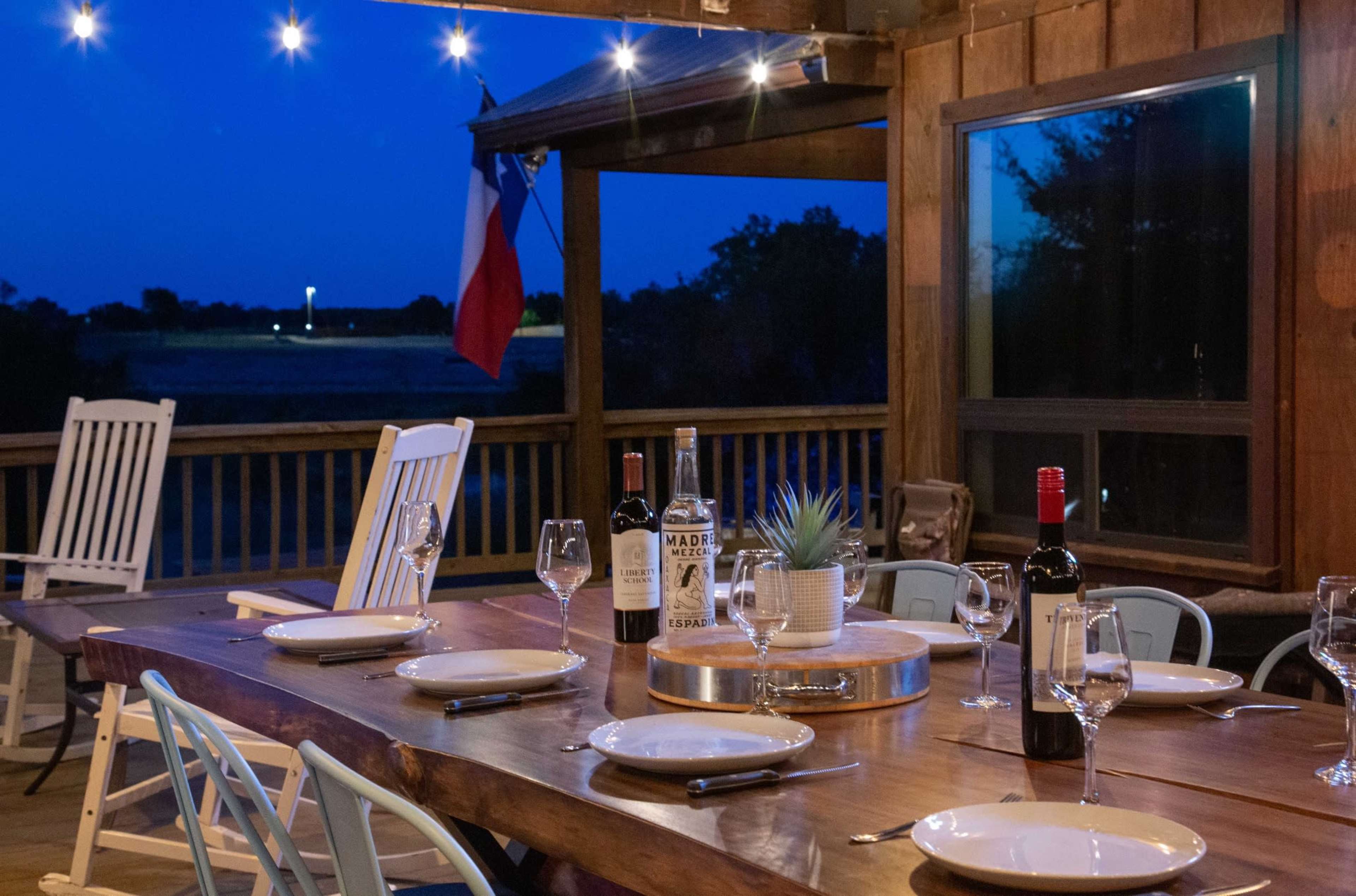 A wooden dining table is set with plates and glasses on a porch at dusk, featuring a Texas flag in the background.