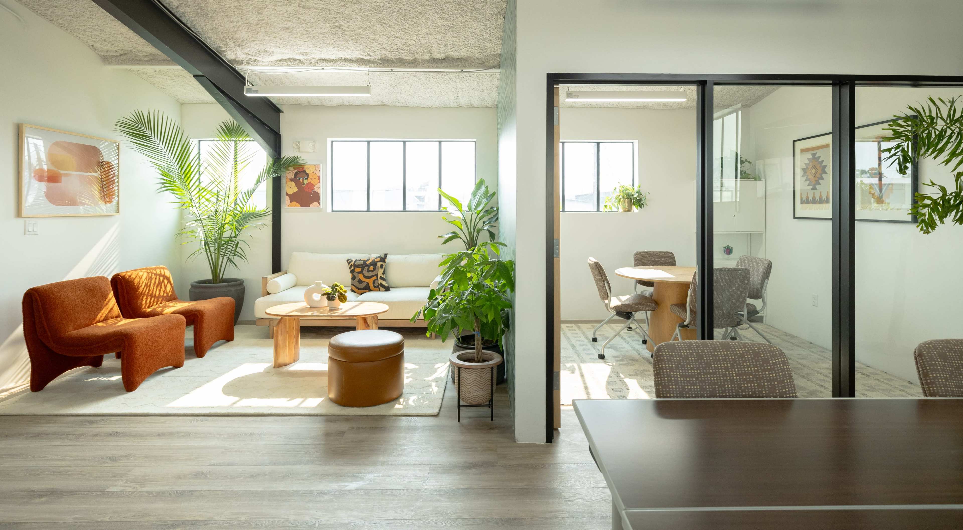 A modern office space divided into a lounge area with orange seating and plants and a meeting area with a circular table and chairs.
