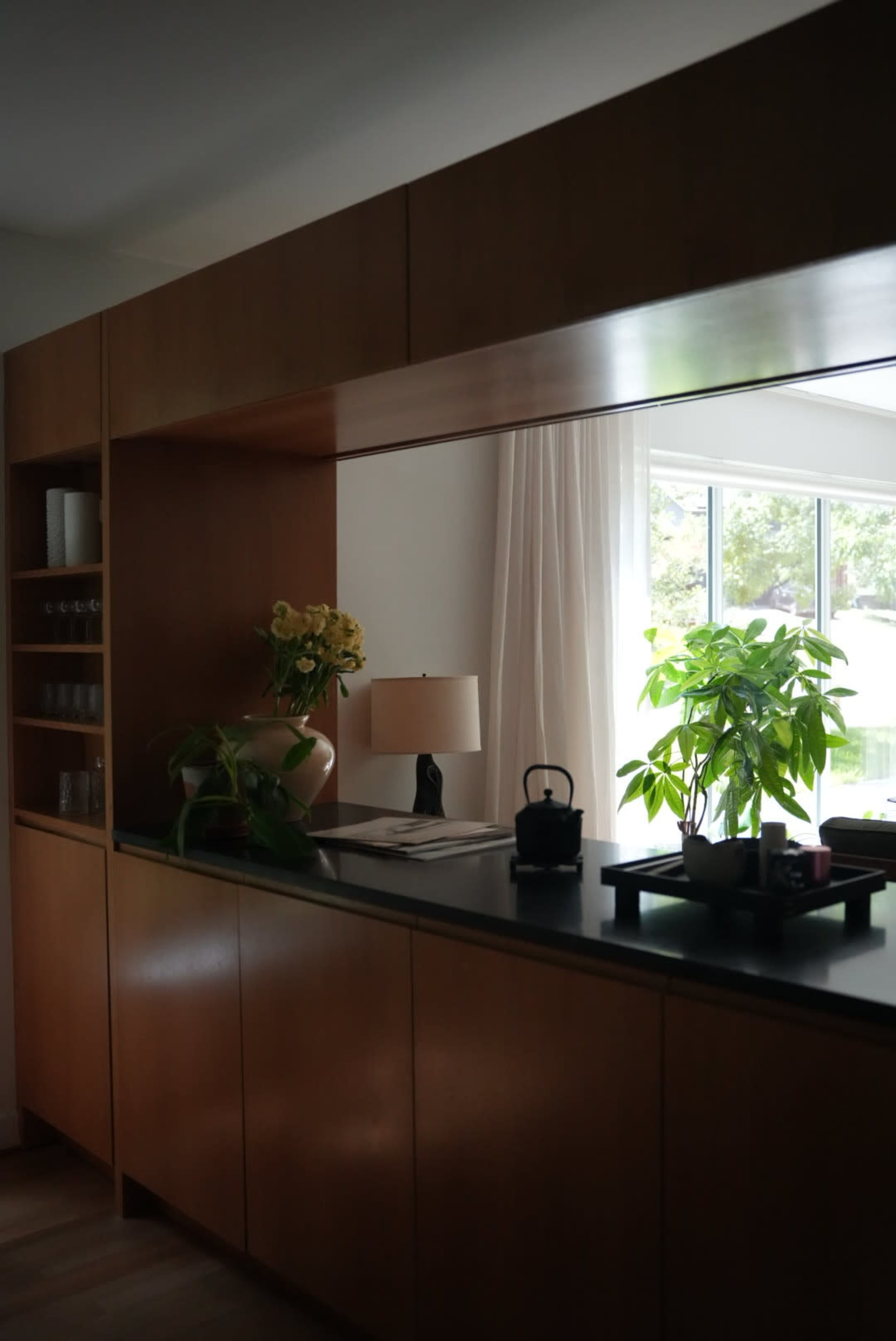 The image shows a modern kitchen with wooden cabinetry, a small lamp, a potted plant, and a view of a window with natural light.
