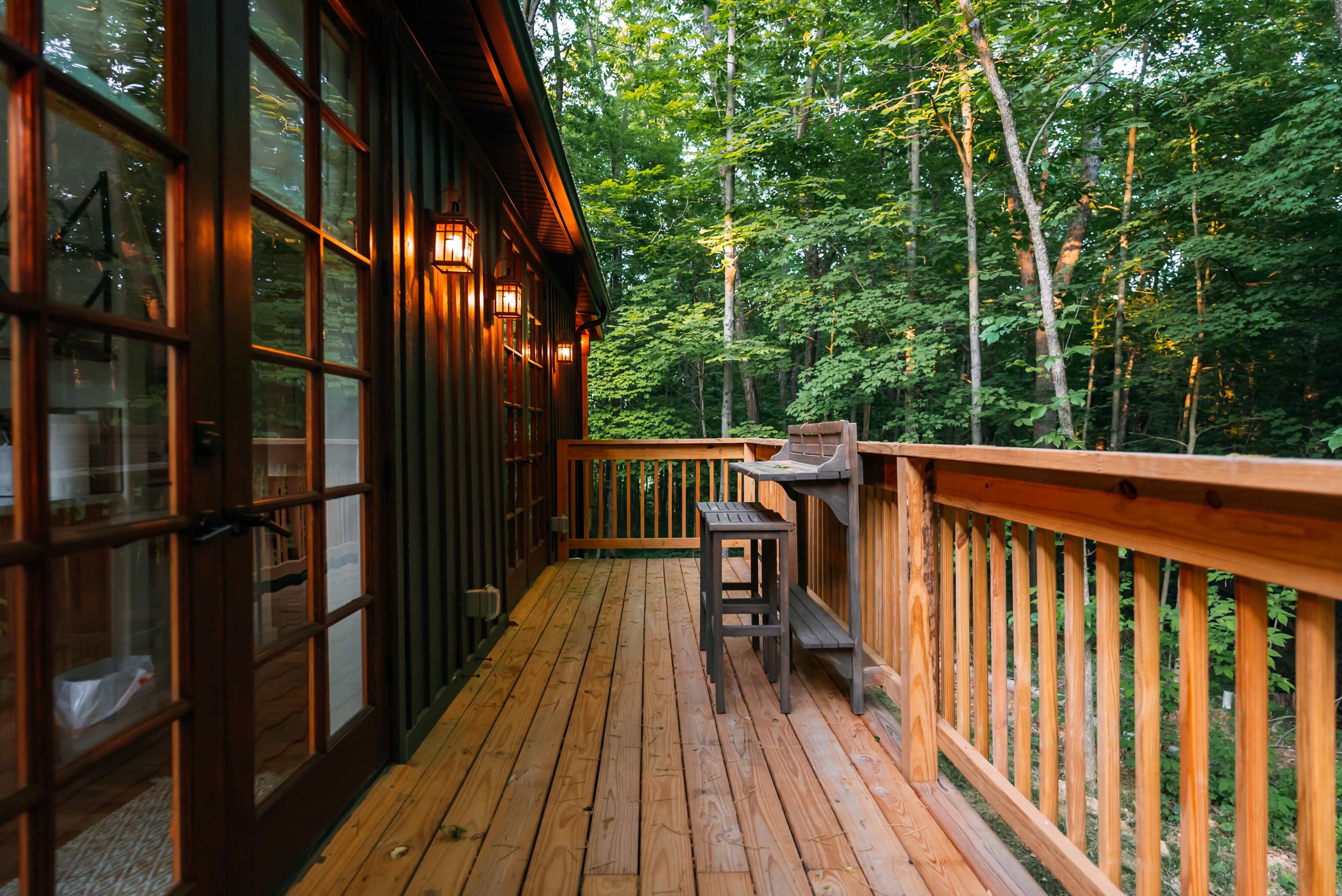 A wooden deck surrounded by trees, with lanterns mounted on the wall and a bar-style table and stools.