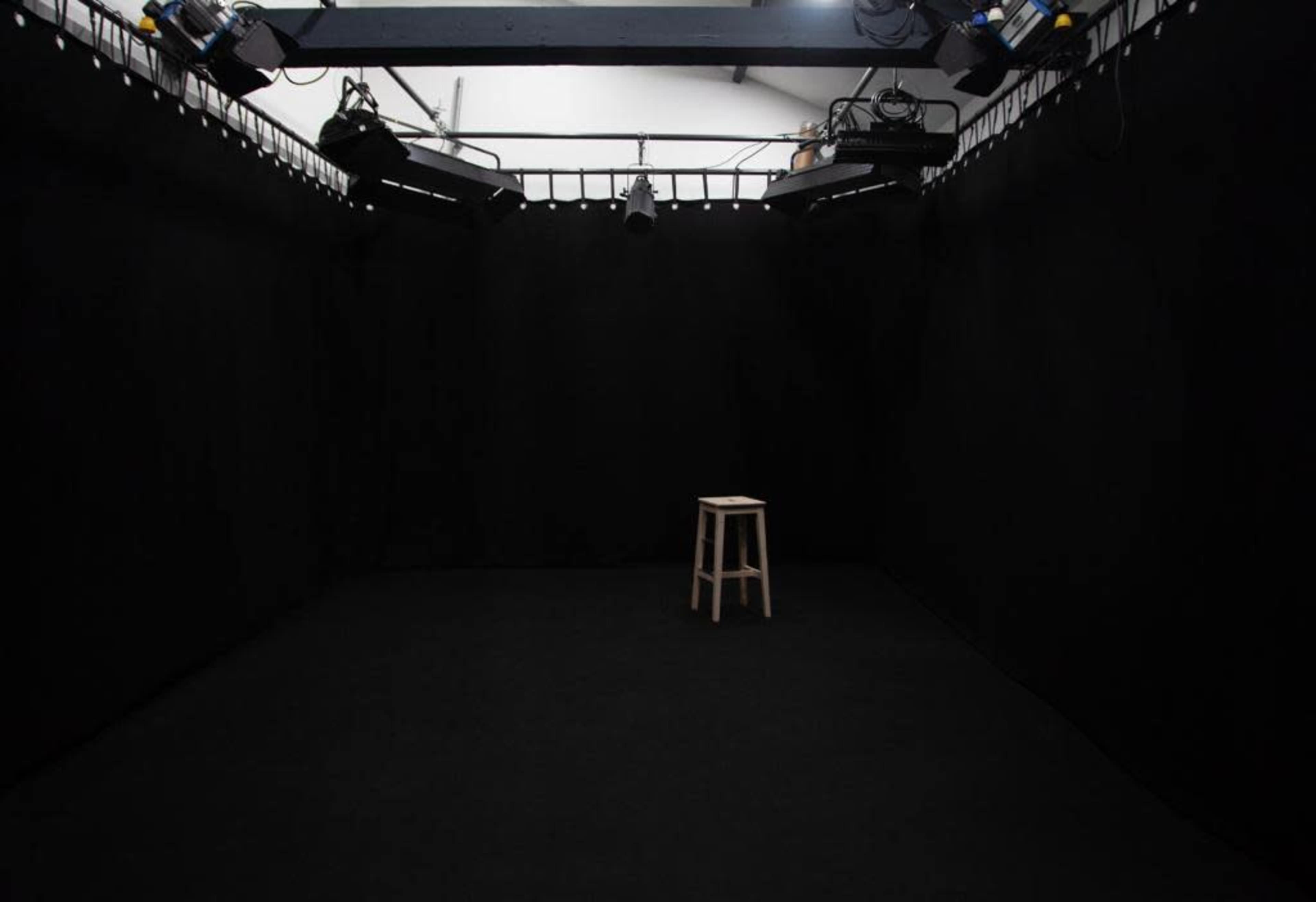 A small, empty studio space with black walls and a single wooden stool in the center, illuminated by overhead lights.