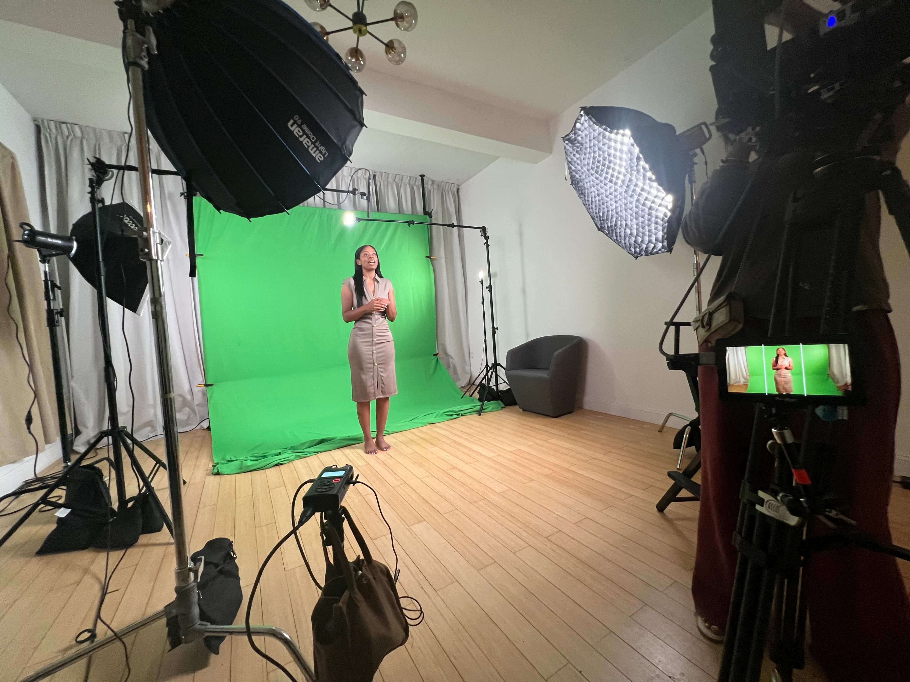 A woman stands in front of a green screen in a studio setting, surrounded by professional lighting and camera equipment.