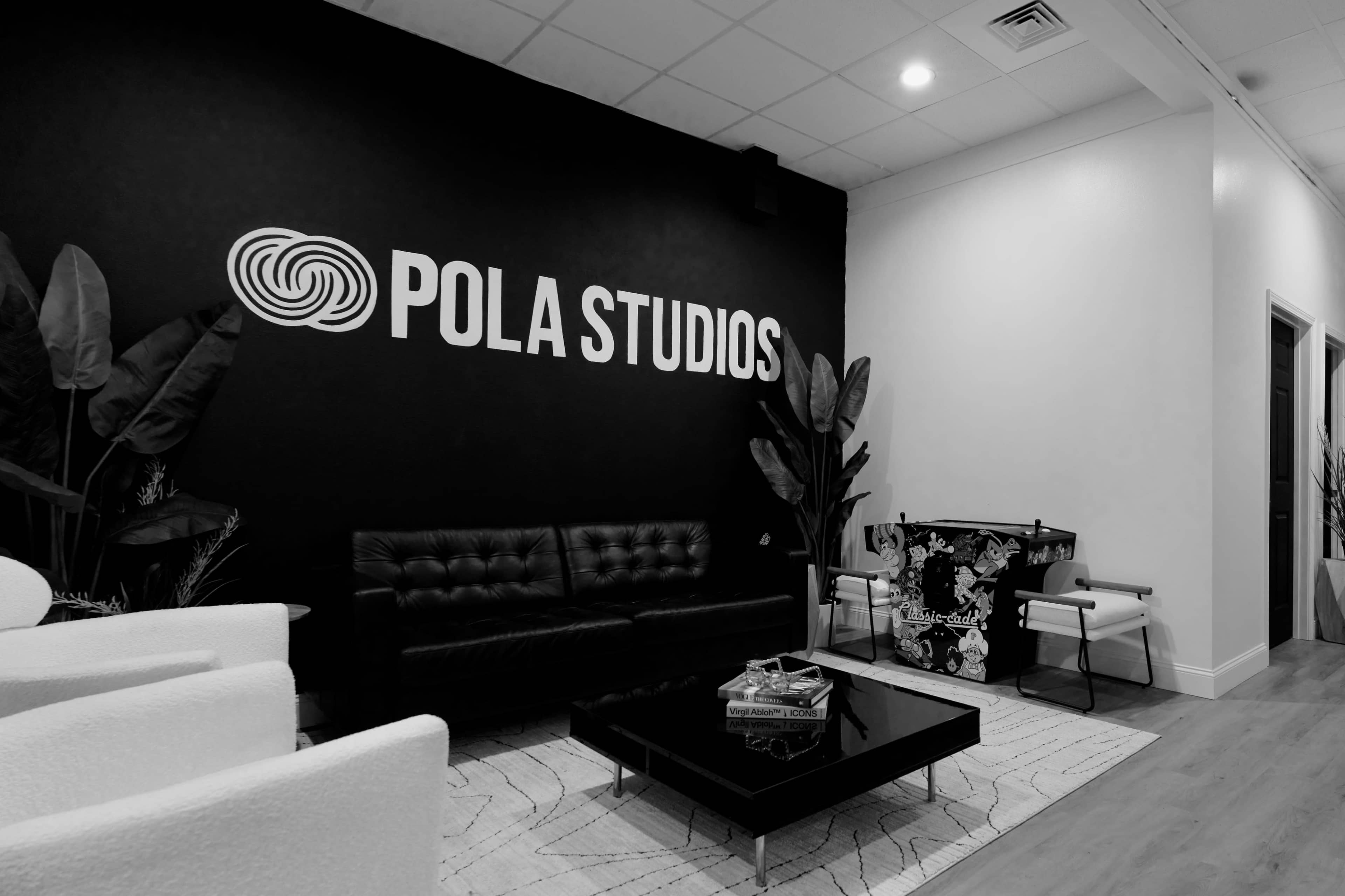 A modern lounge area featuring a black wall with "POLA STUDIOS" prominently displayed, a black leather sofa, a glass coffee table, and decorative plants.