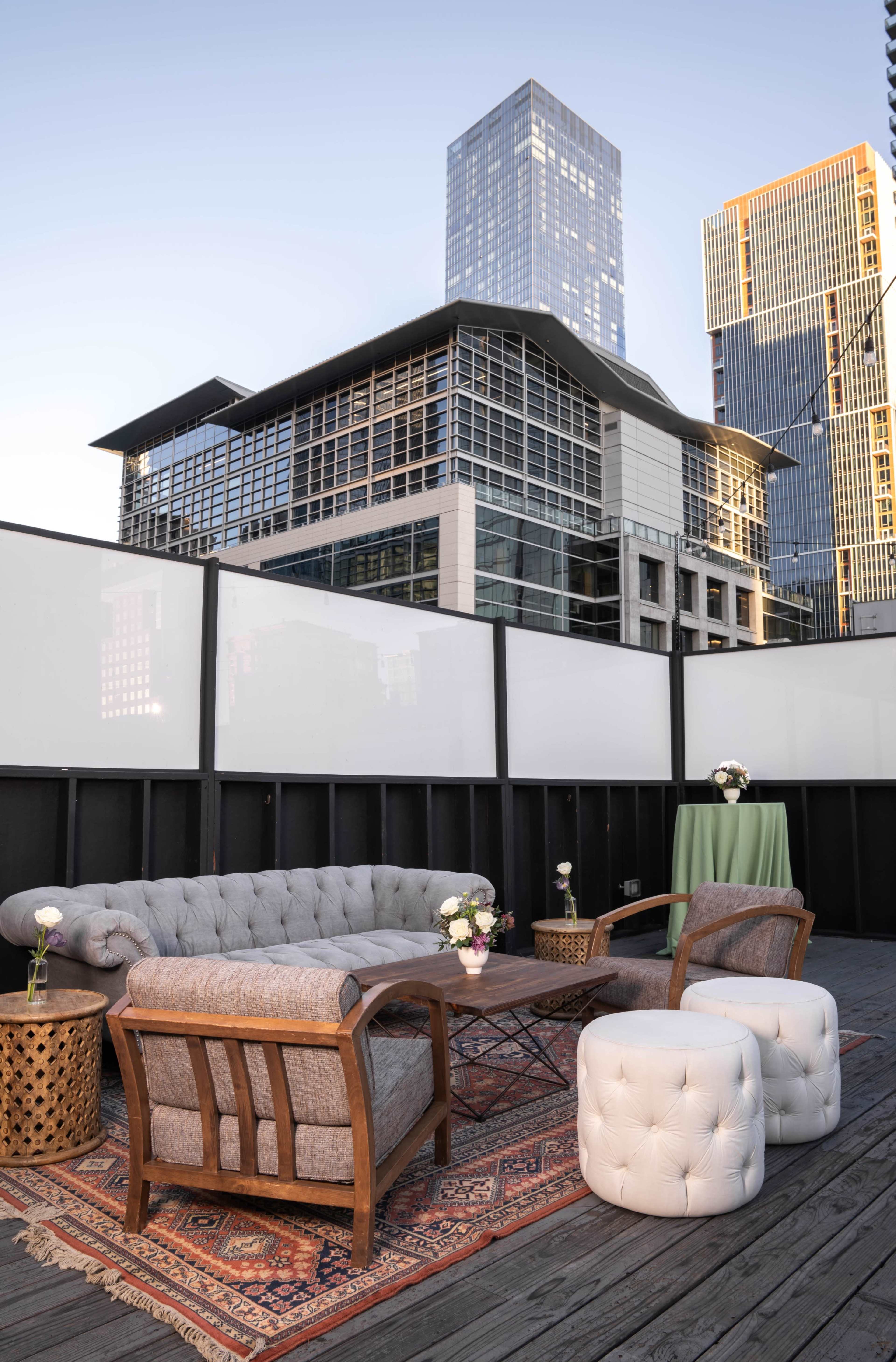 A rooftop lounge area features a tufted sofa, two wooden chairs, a small table, and tall buildings in the background.