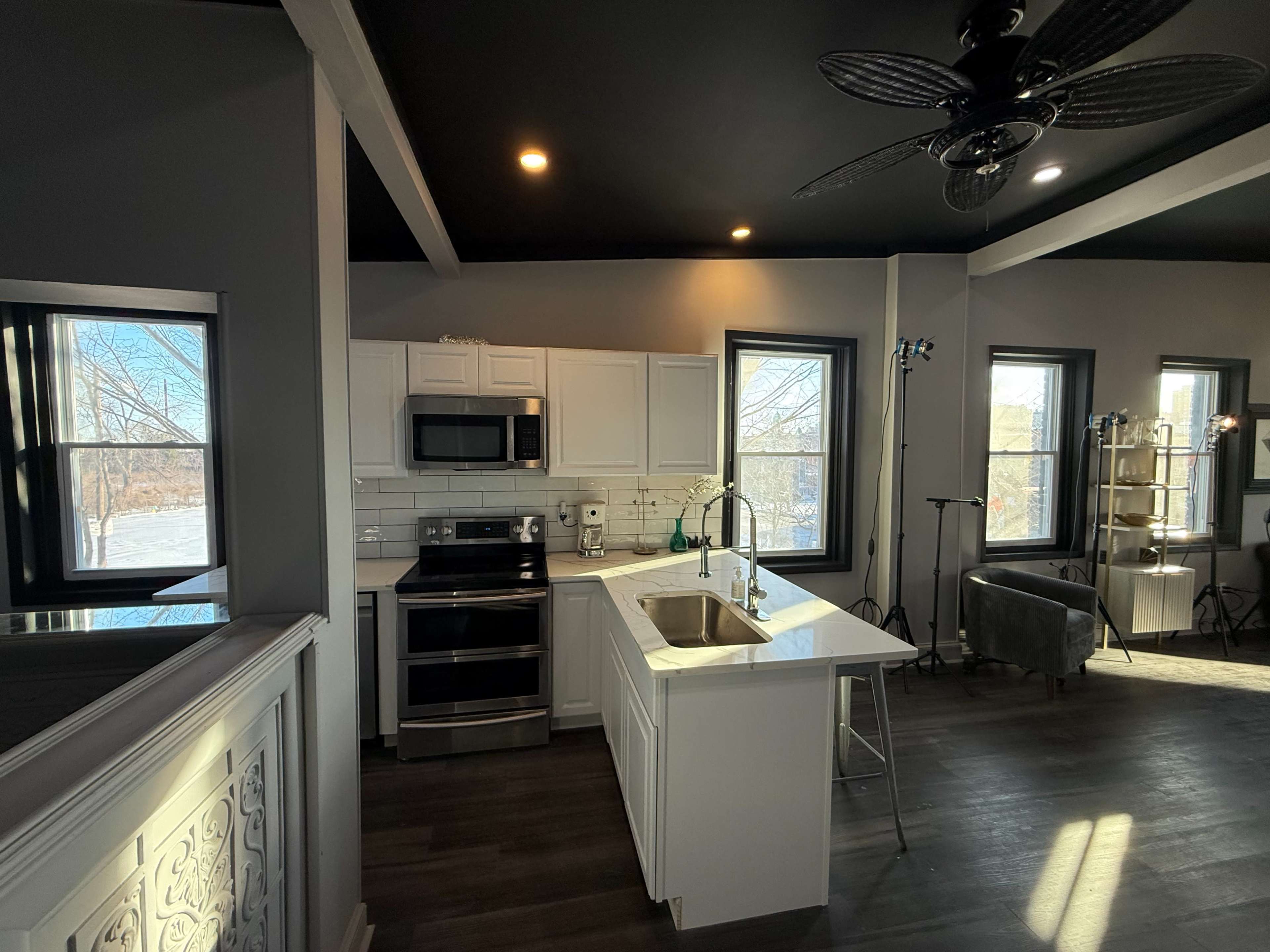 The image shows a modern kitchen featuring white cabinetry, stainless steel appliances, and a sink with a view of a spacious room filled with natural light.