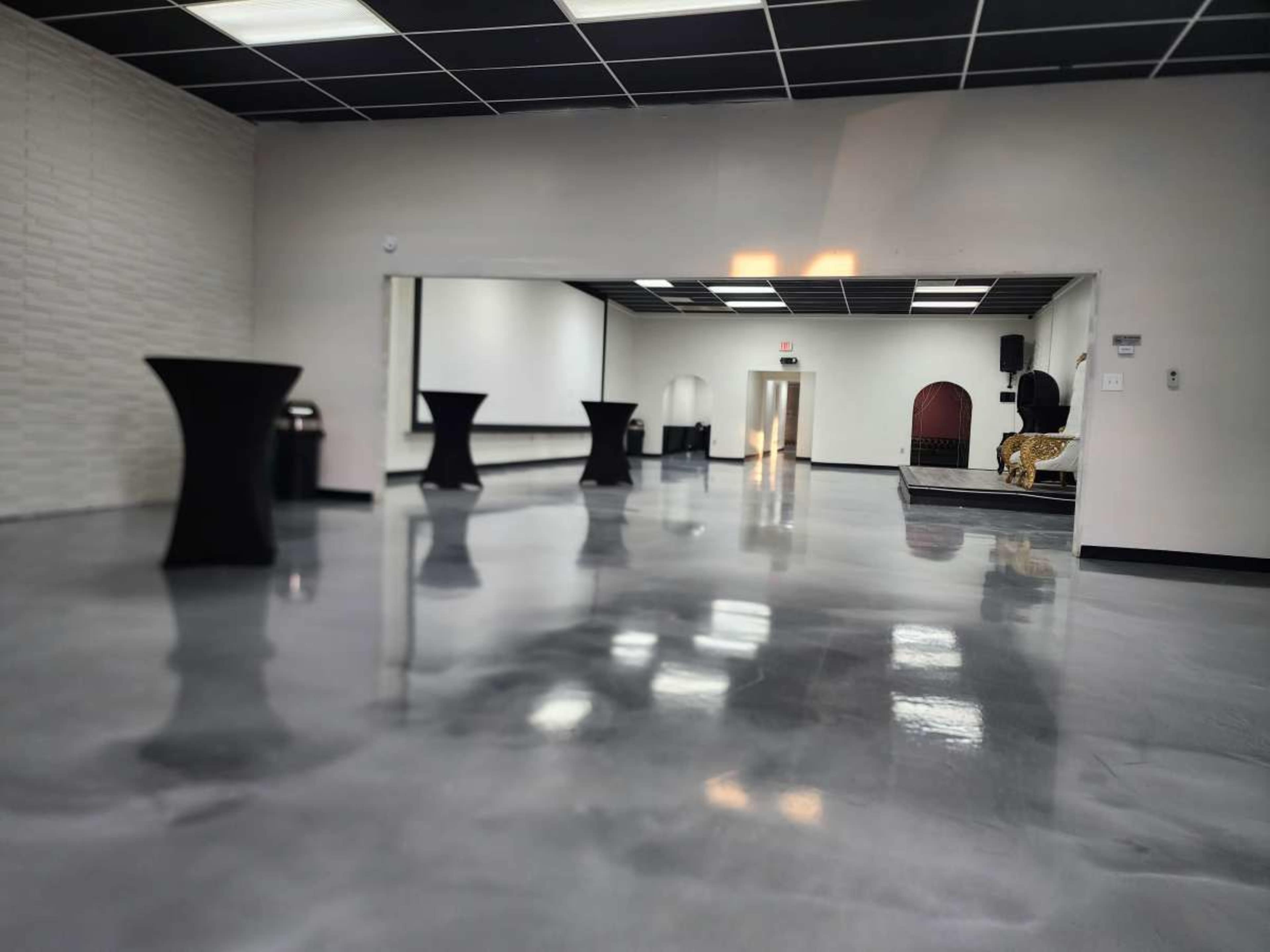 The image shows a spacious, empty room with polished concrete floors, black cocktail tables, and a small stage in the background.