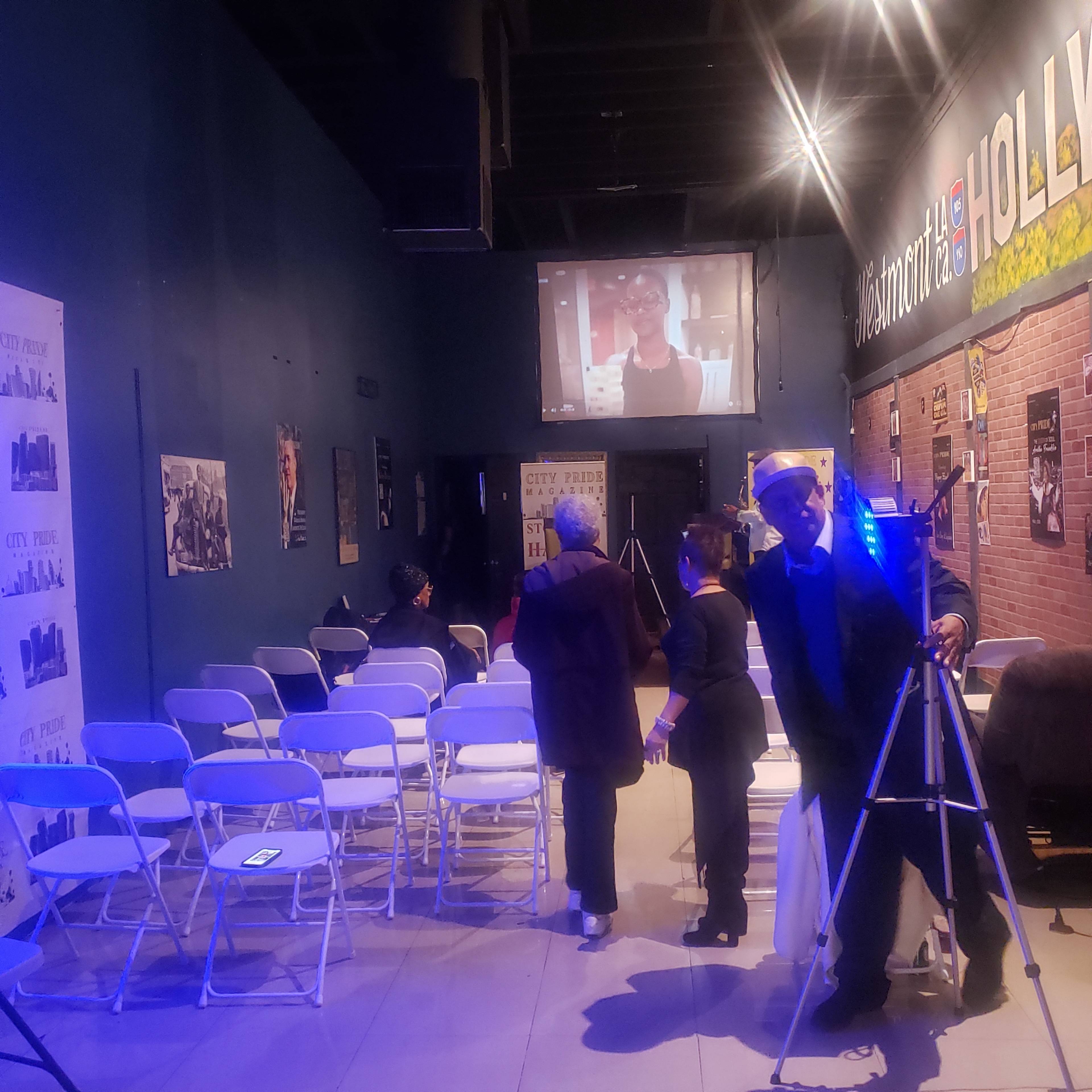 A small audience gathers in a dimly lit room with folding chairs, while a screen displays a video featuring a woman.