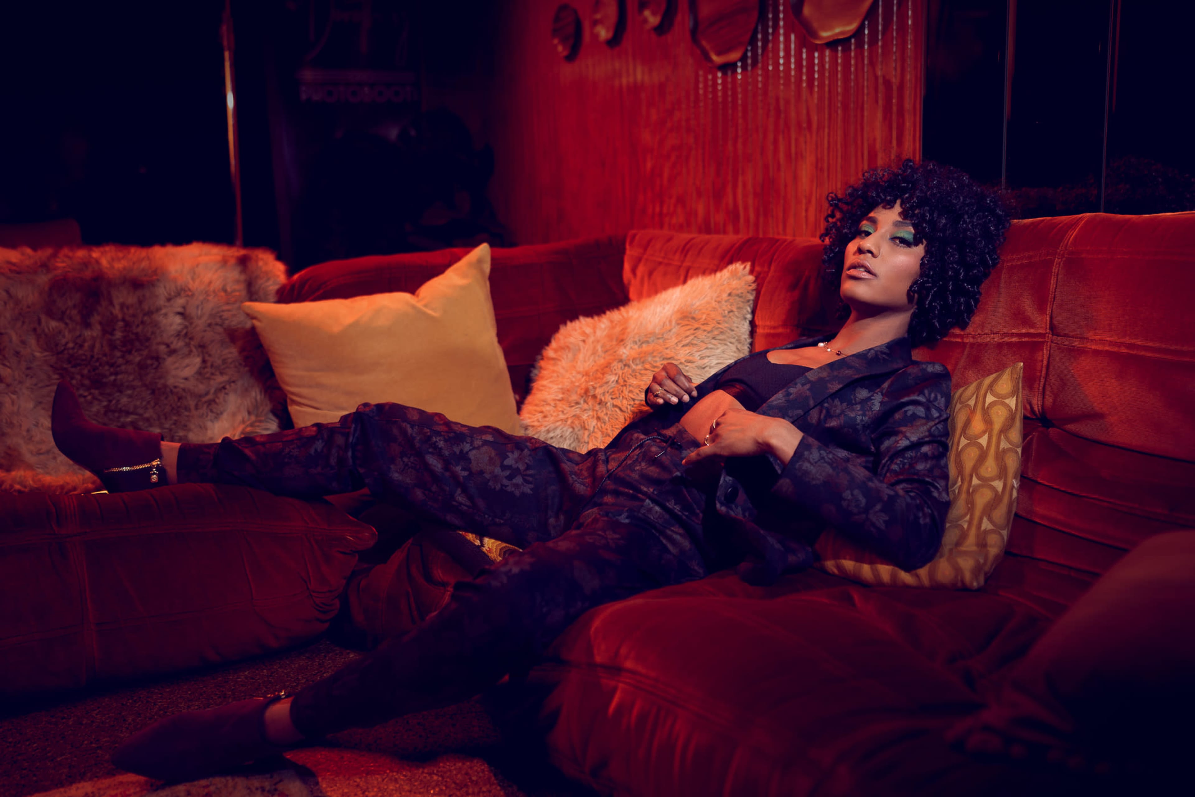 A person with curly hair lounges on a red sofa adorned with decorative pillows in a dimly lit room.