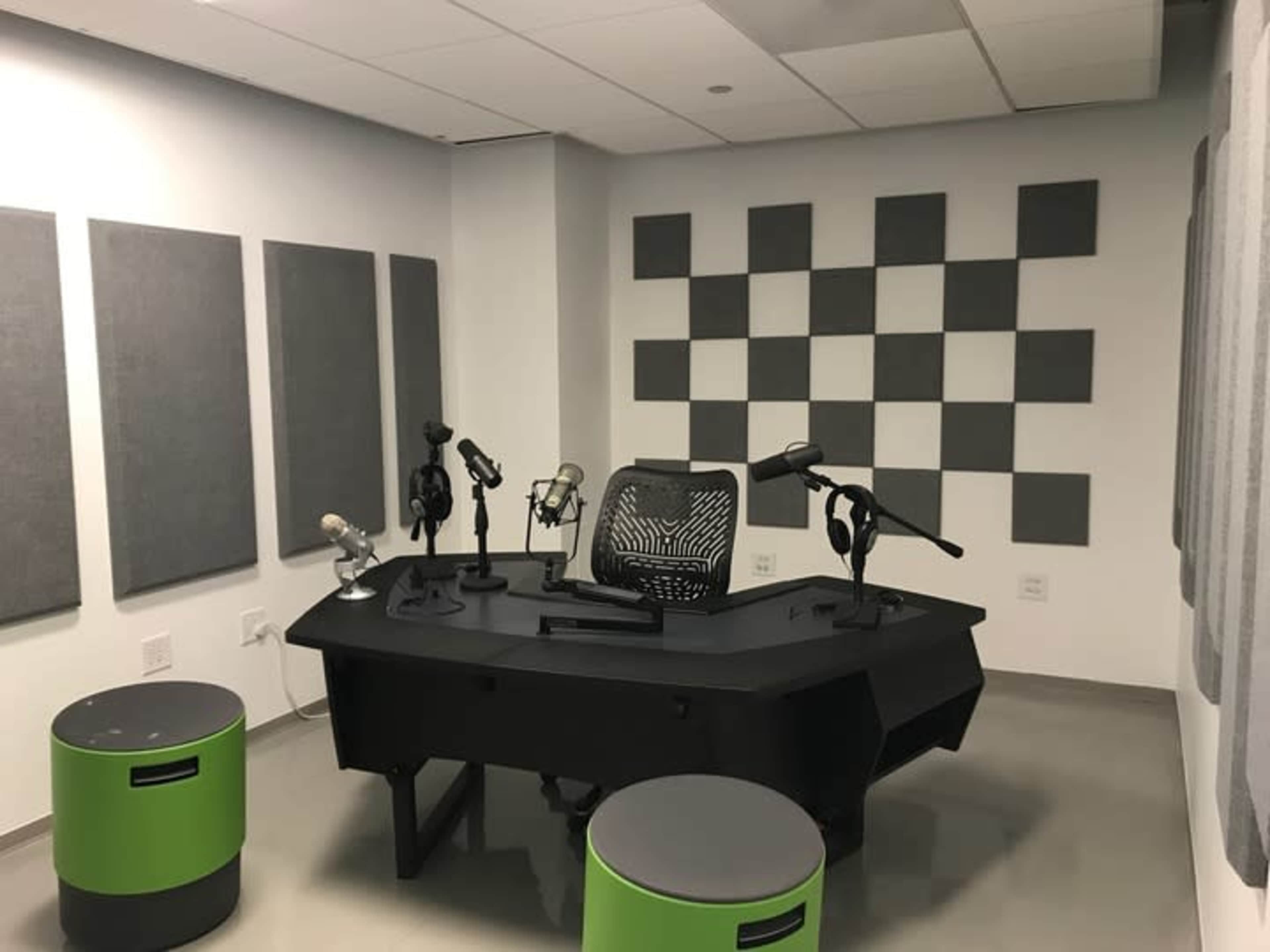 The image shows a modern recording studio with a black desk, multiple microphones, and sound-absorbing panels on the walls.