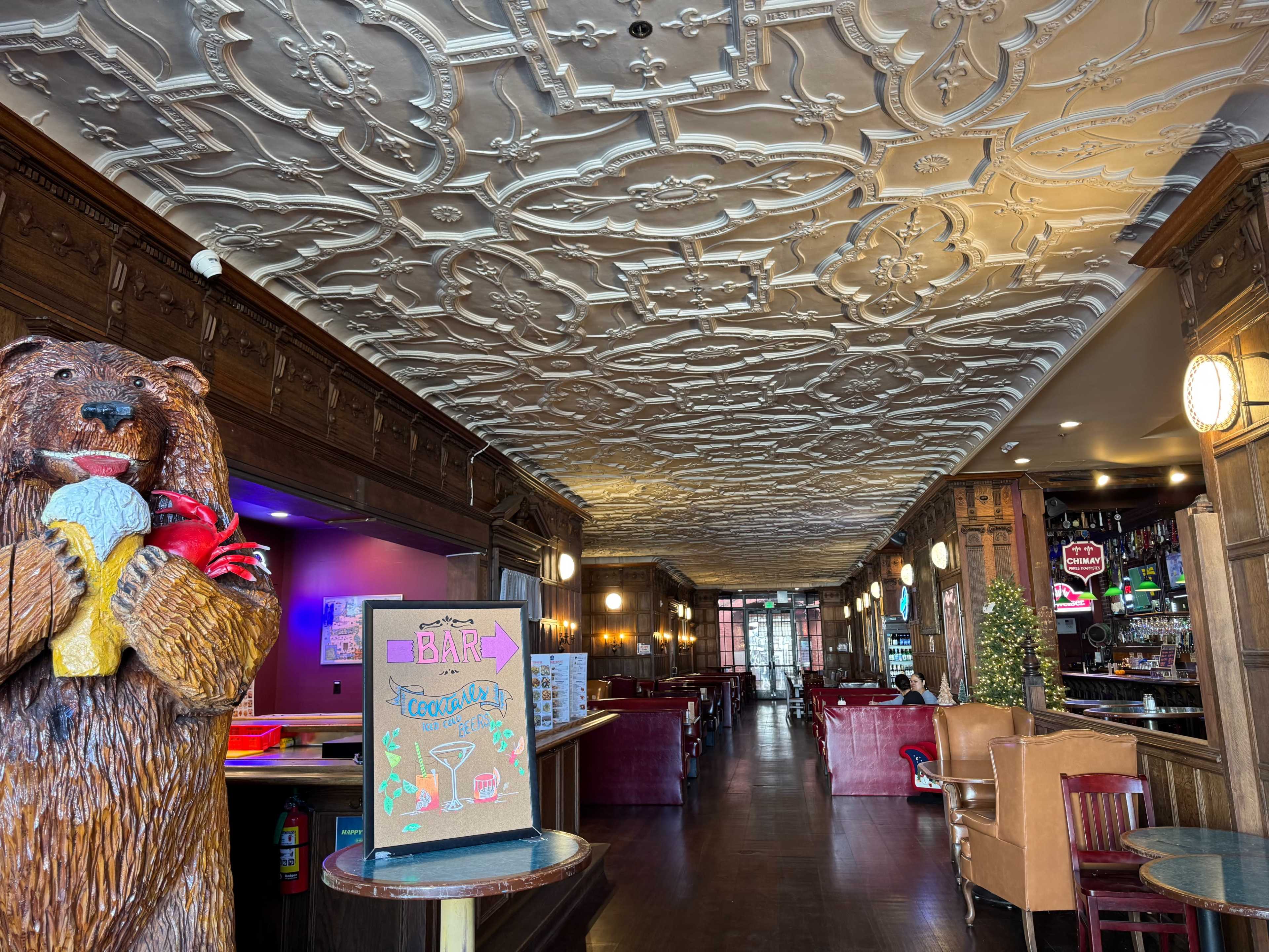The image depicts a long corridor of a vintage-styled bar or restaurant, featuring decorative ceiling patterns, wooden paneling, and a bear statue holding ice cream near the entrance.
