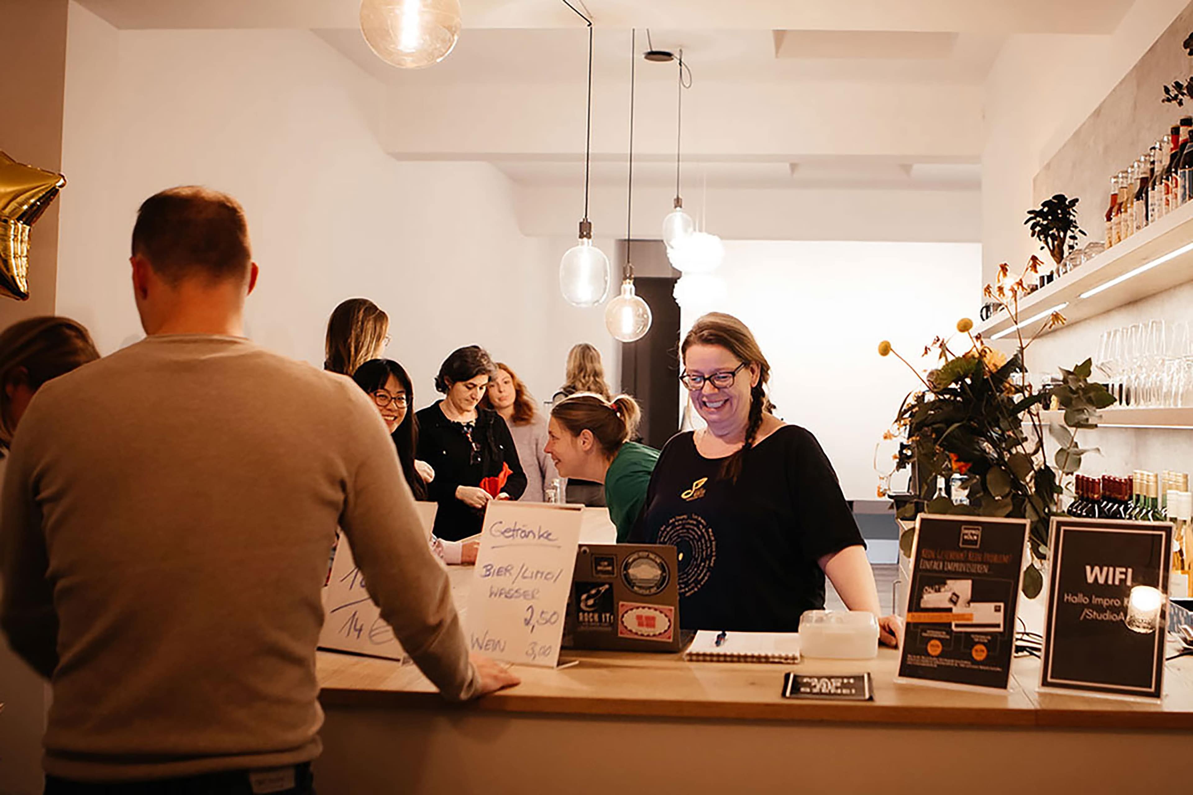 A busy café scene shows a group of people socializing at a counter while a staff member assists a customer.