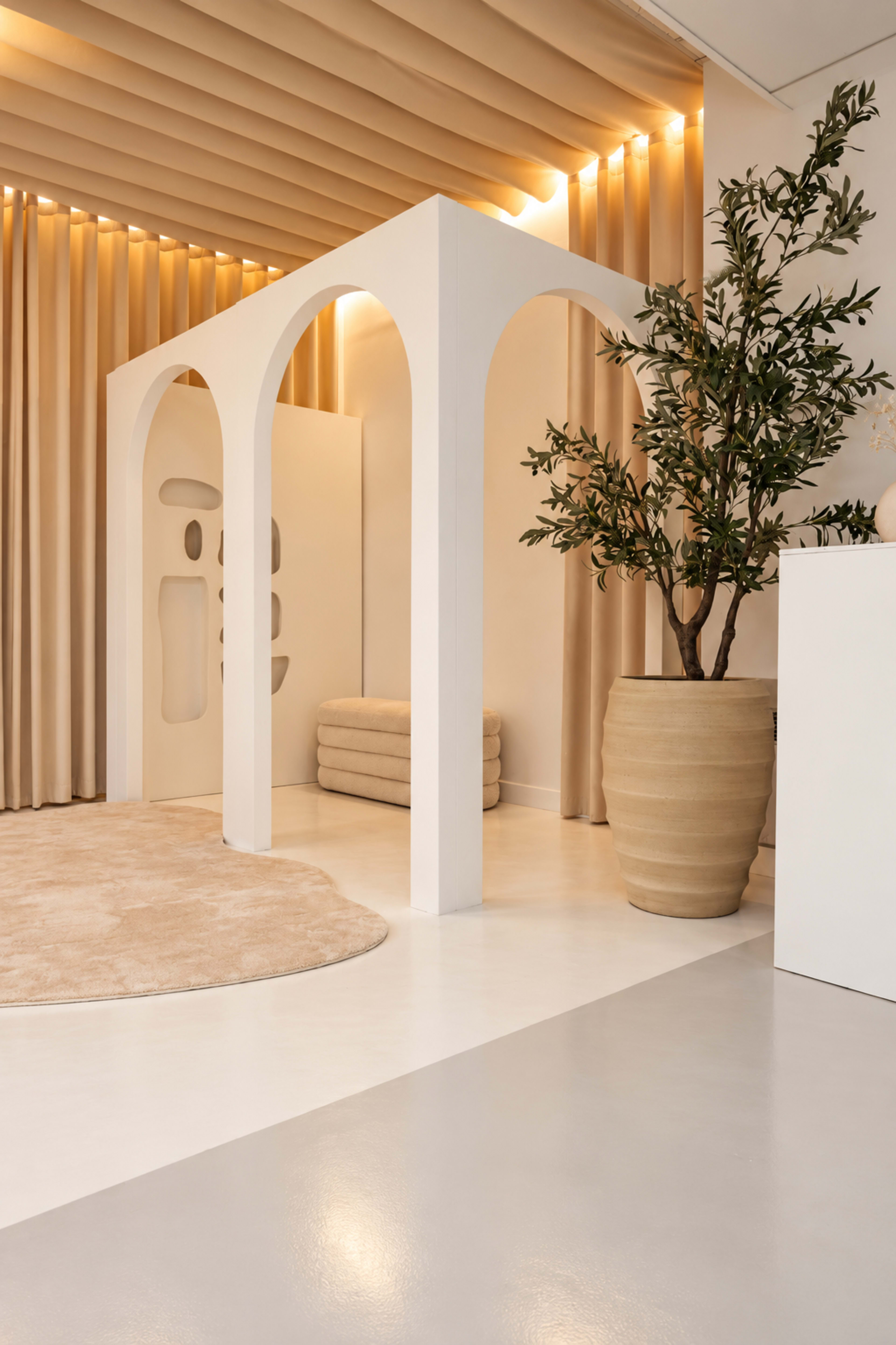 The image features a modern interior space with a white archway structure, a large plant in a beige pot, and soft textures in neutral colors.