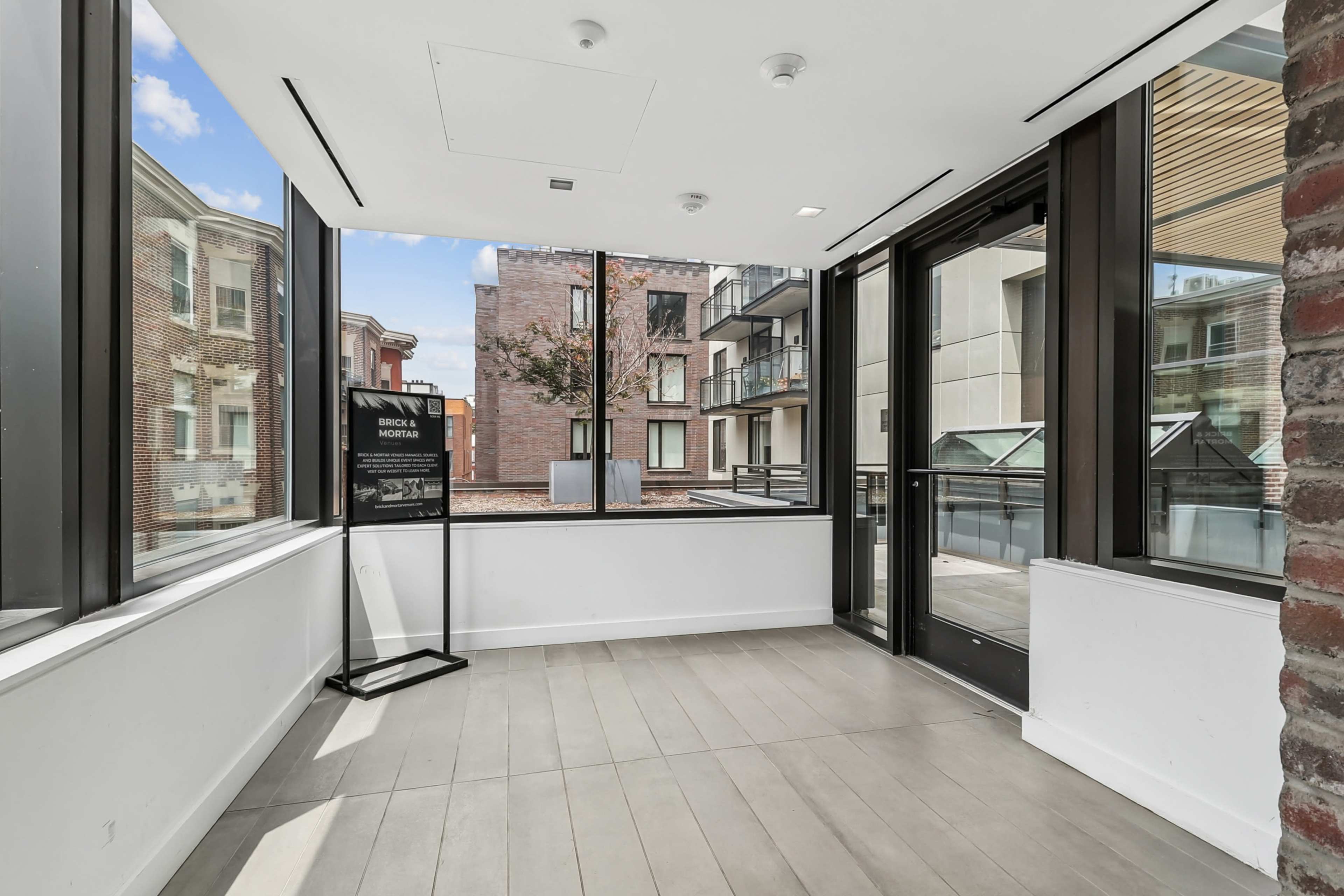 The image depicts a modern, sunlit interior space with large windows overlooking a courtyard and adjacent buildings.