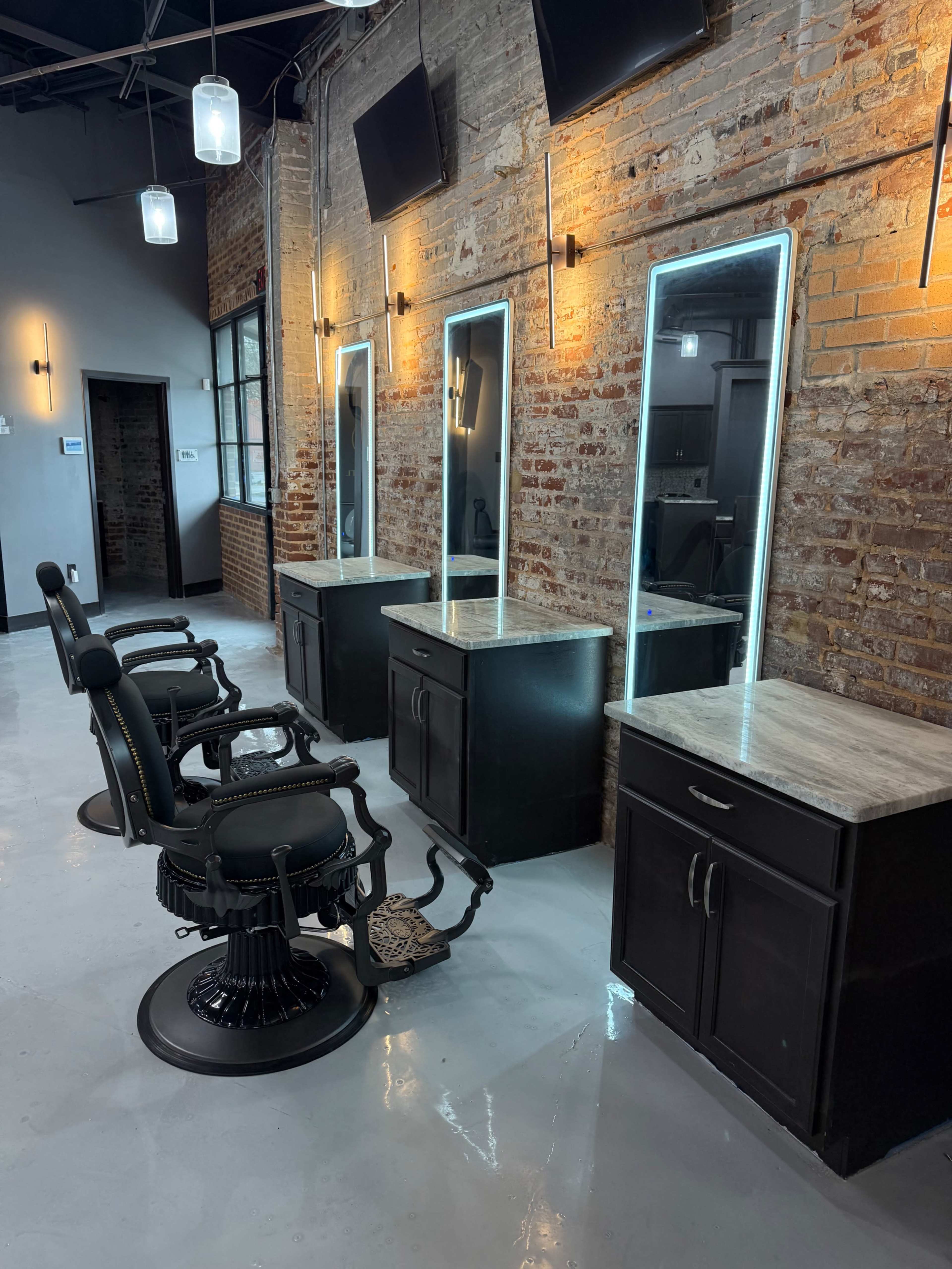 The image shows a modern salon interior with four styling stations featuring mirrors and granite countertops, alongside black barber chairs on a polished concrete floor.