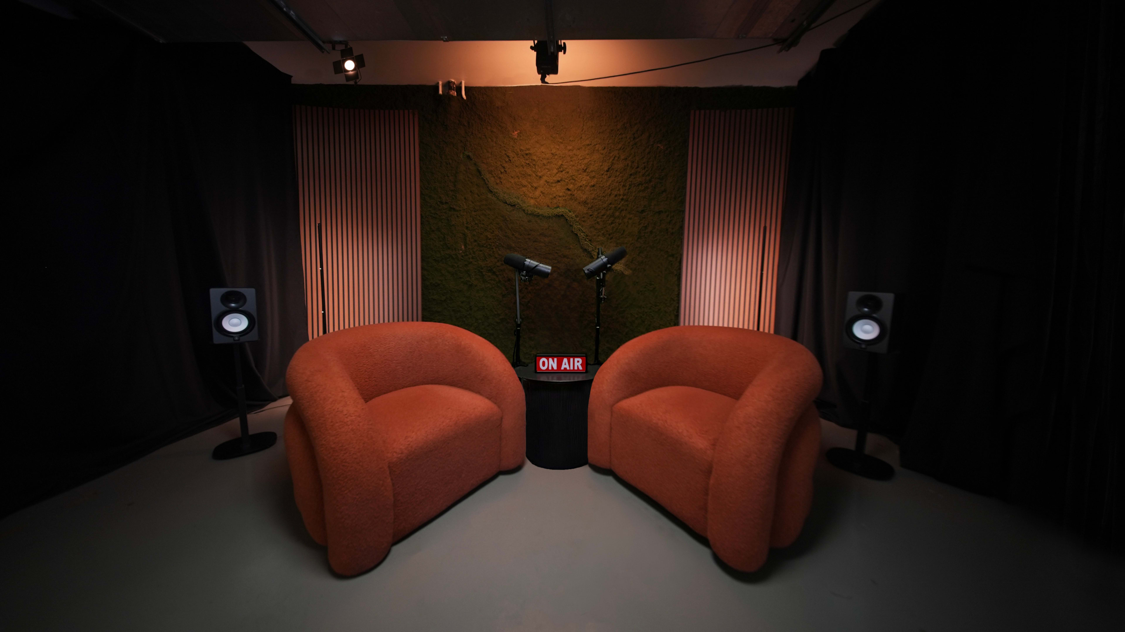 The image features a cozy podcast studio setup with two orange armchairs facing a small table that has a "ON AIR" sign, flanked by two microphones and speakers against a dark backdrop.