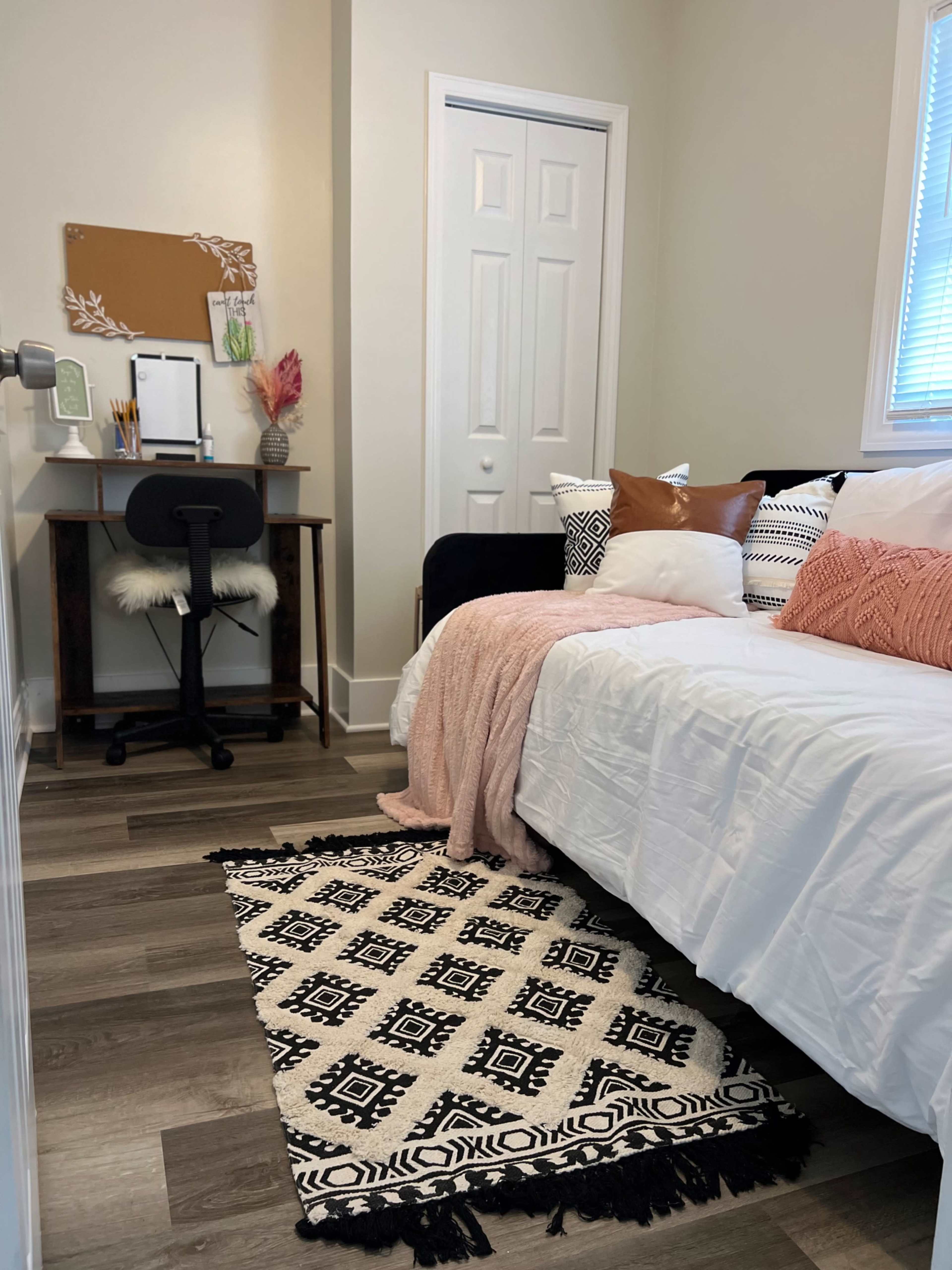 A small, organized bedroom featuring a bed with decorative pillows, a desk with a chair, and a patterned rug on the floor.