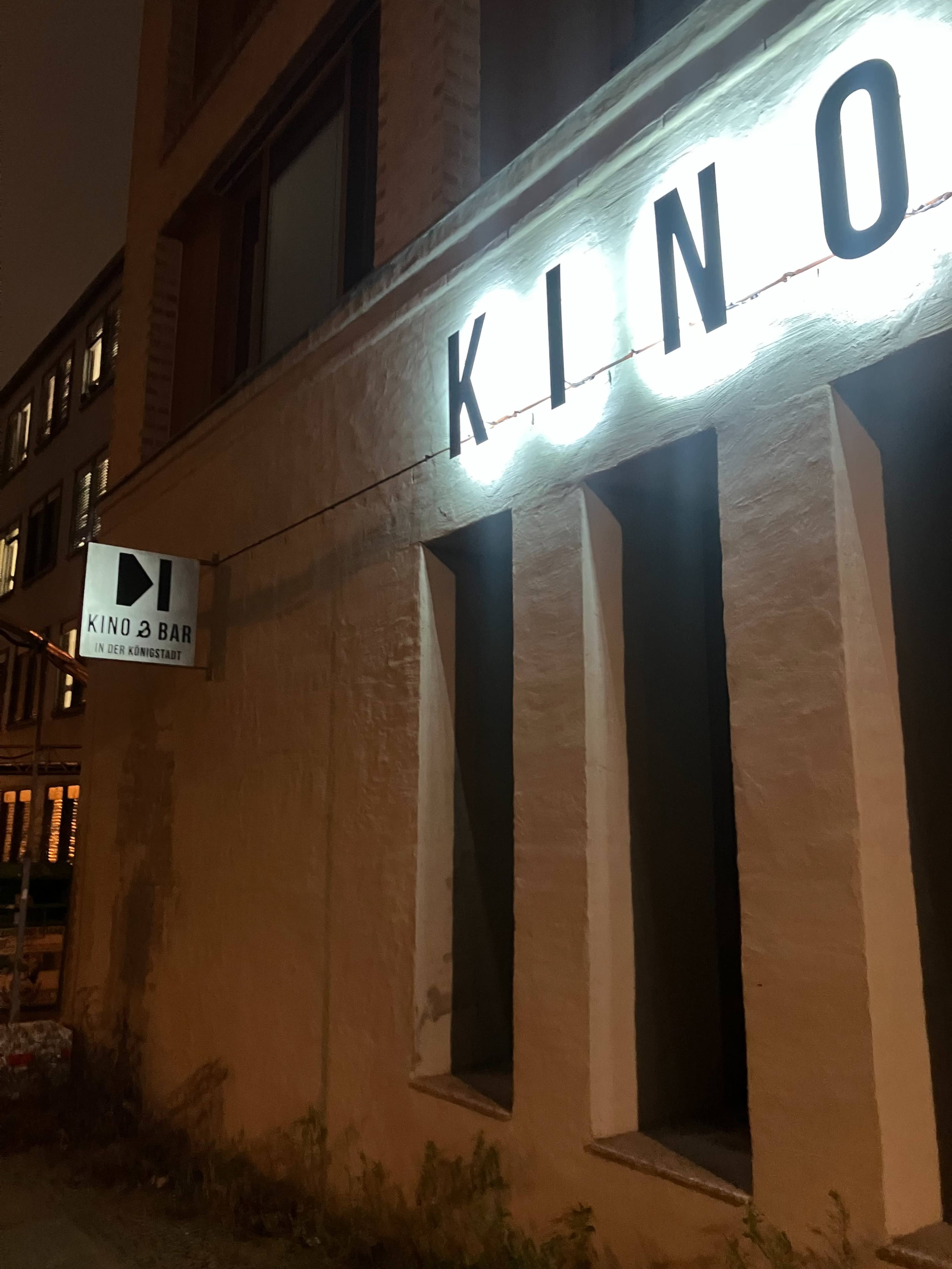The image shows the illuminated sign of a venue called "KINO" on the side of a building, with a smaller sign indicating a bar.