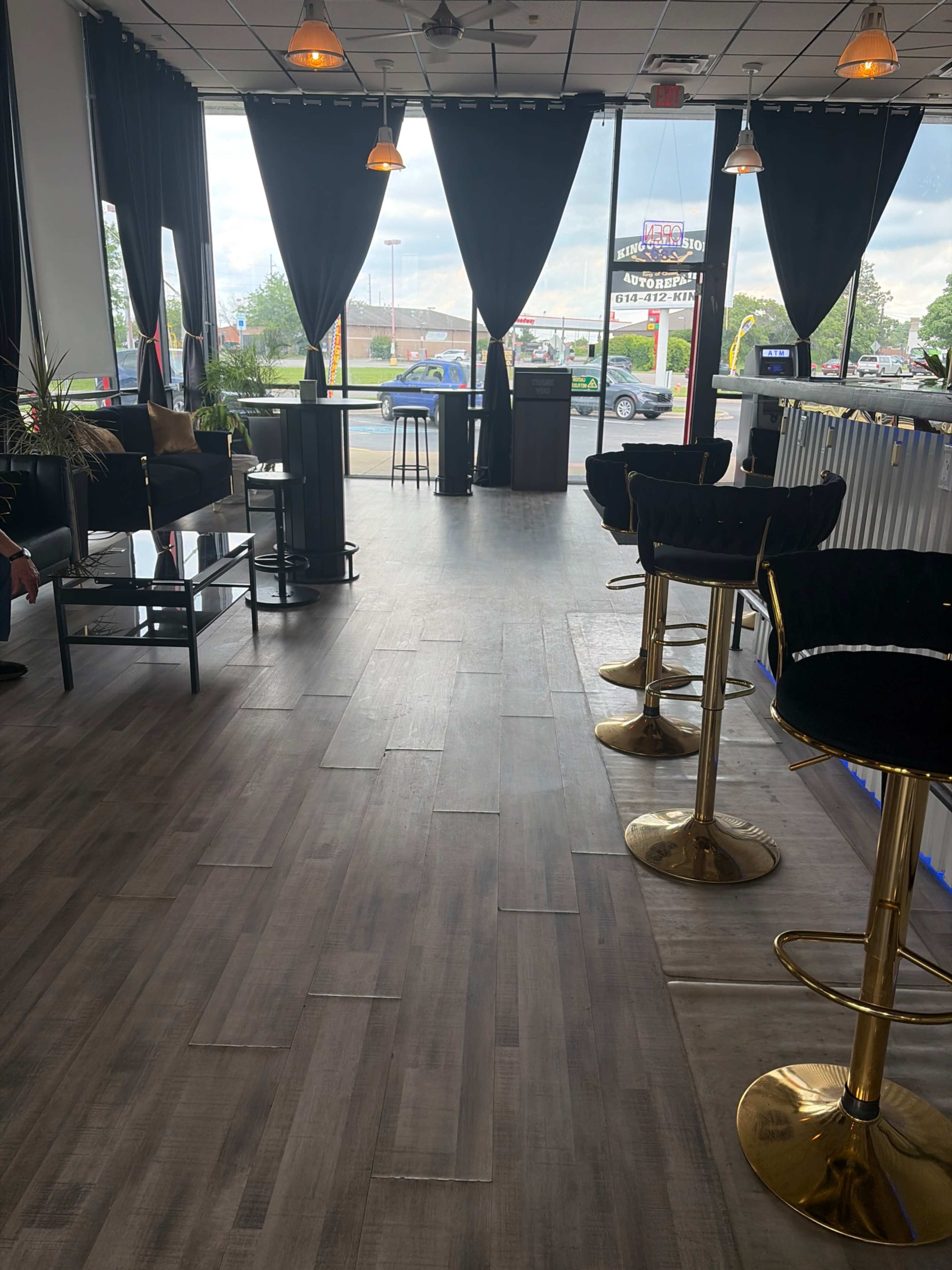 The image shows an interior space with wood-like flooring, black curtains, bar stools, and small tables, illuminated by natural light through large windows.