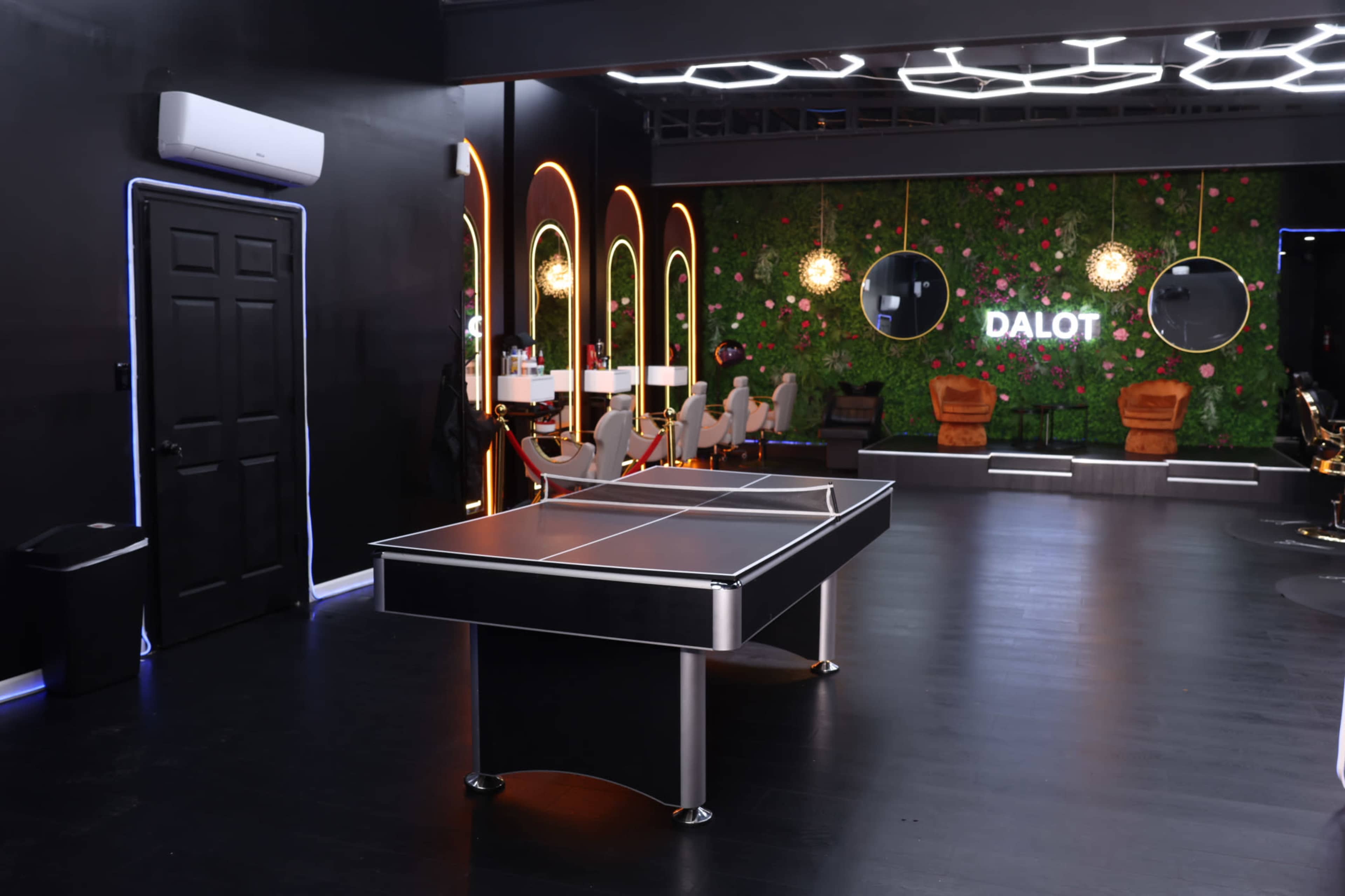 The image shows a modern lounge area with a ping pong table in the foreground, stylish seating, and decorative walls featuring greenery and illuminated mirrors in the background.
