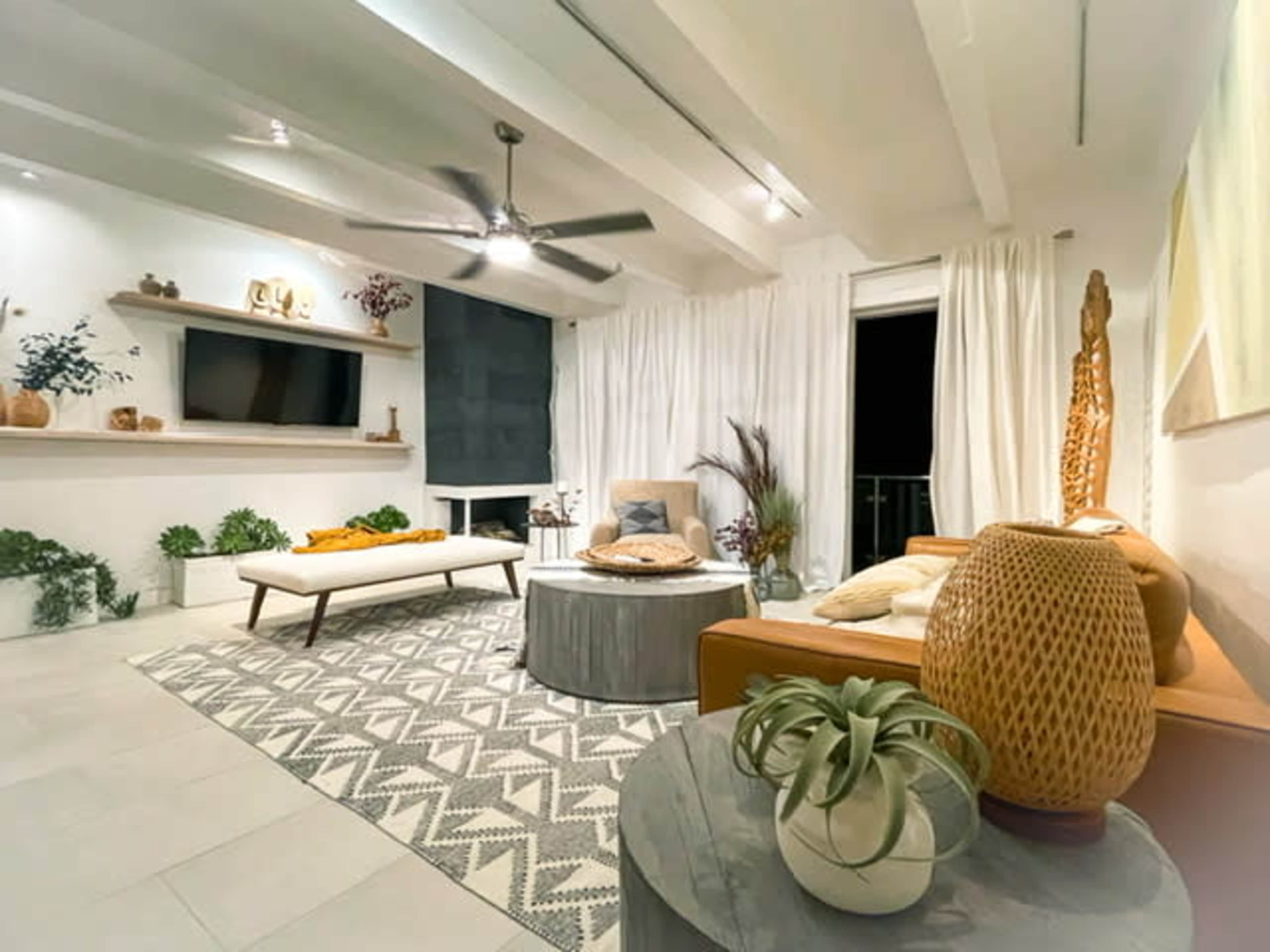 A modern living room featuring a light-colored sofa, a coffee table, a ceiling fan, and large windows covered with sheer curtains.